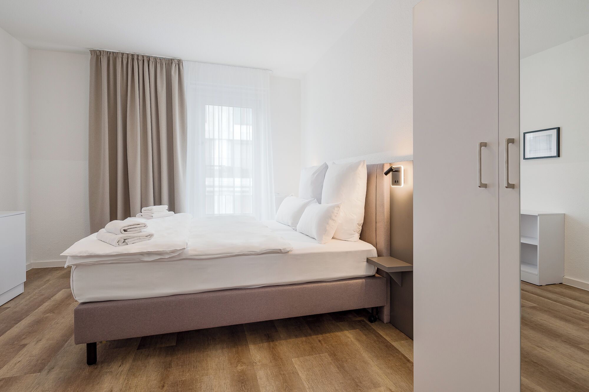 A neatly made bed with white linens, a bedside table with a lamp, and a window with sheer curtains.
