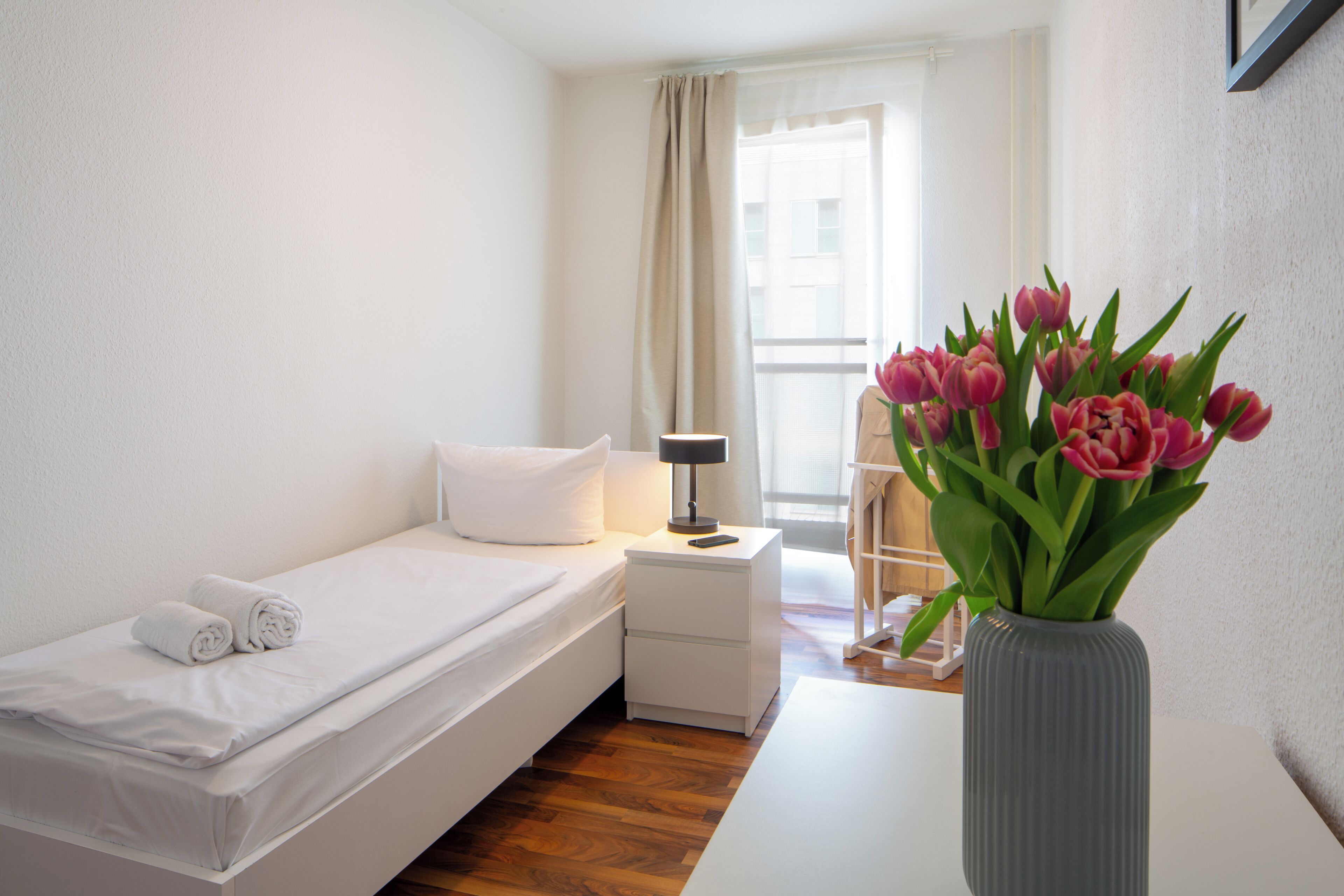 Ein Schlafzimmer mit Bett, Nachttisch, Lampe, Blumenvase und einem Fenster mit Vorhängen.