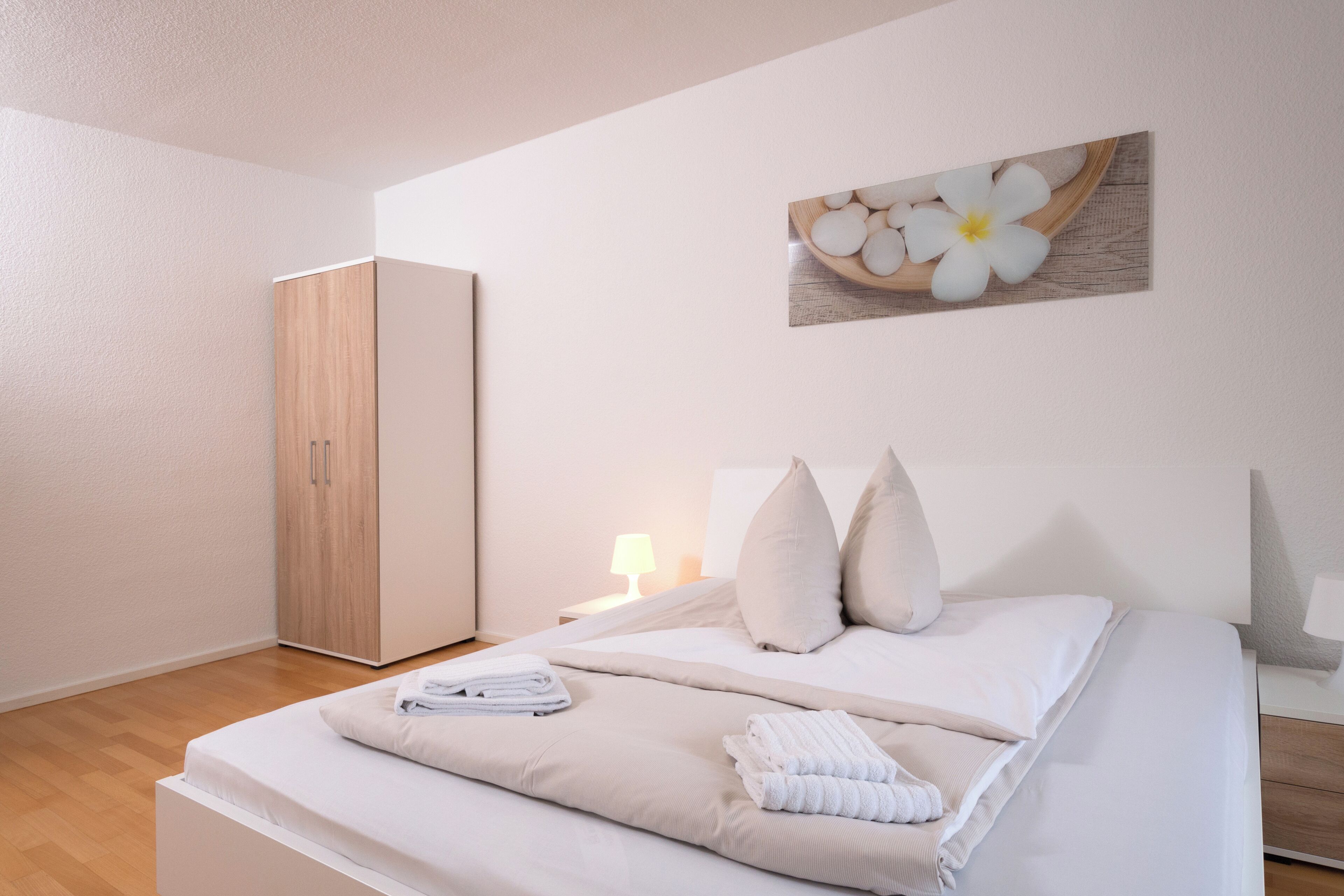 A bedroom with a bed, pillows, a nightstand, a wooden wardrobe, and a wall-mounted picture of flowers.