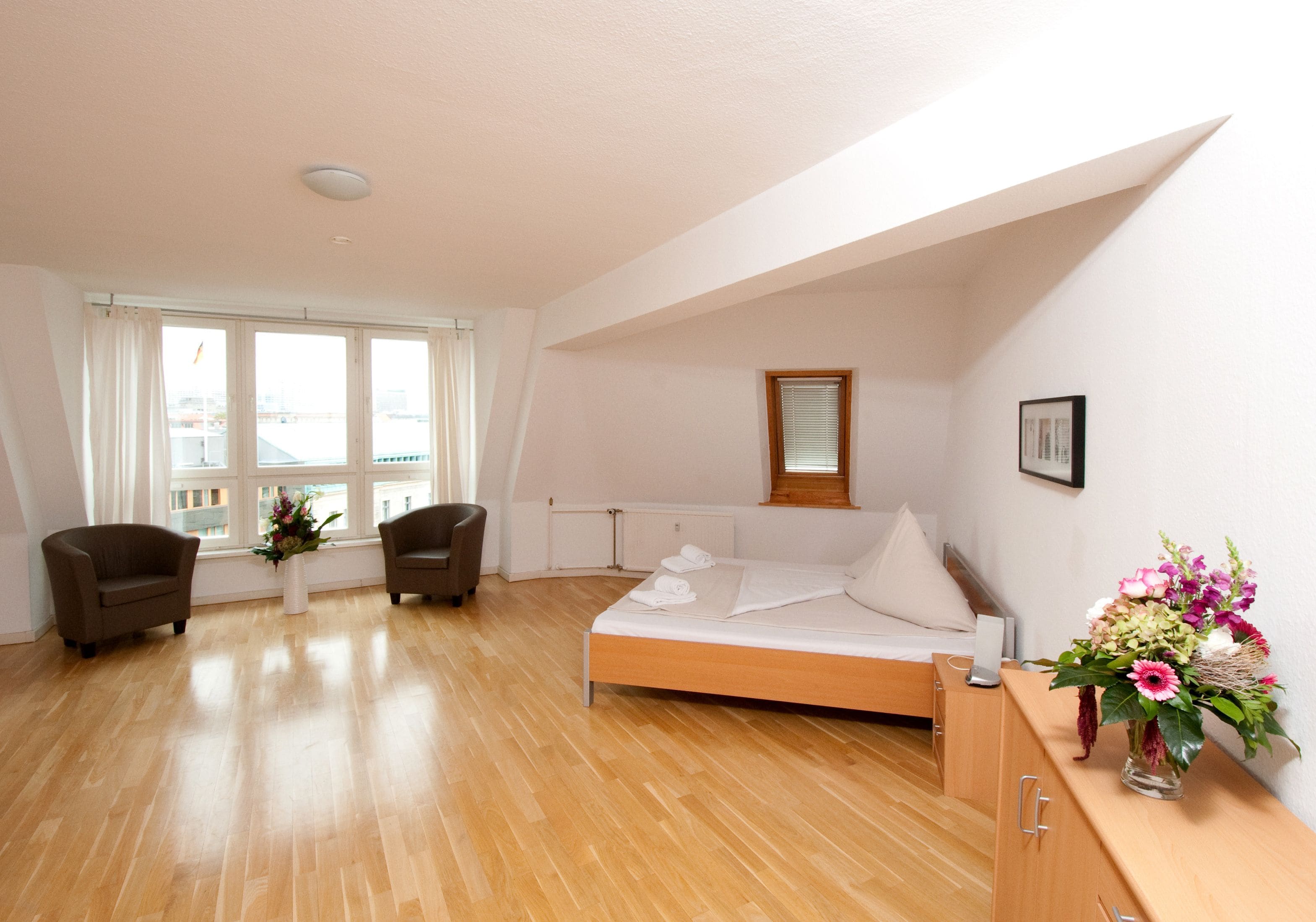 Camera d'albergo con pavimento in legno, un letto con lenzuola bianche, due poltrone, un tavolino con un vaso di fiori e una finestra con tende.