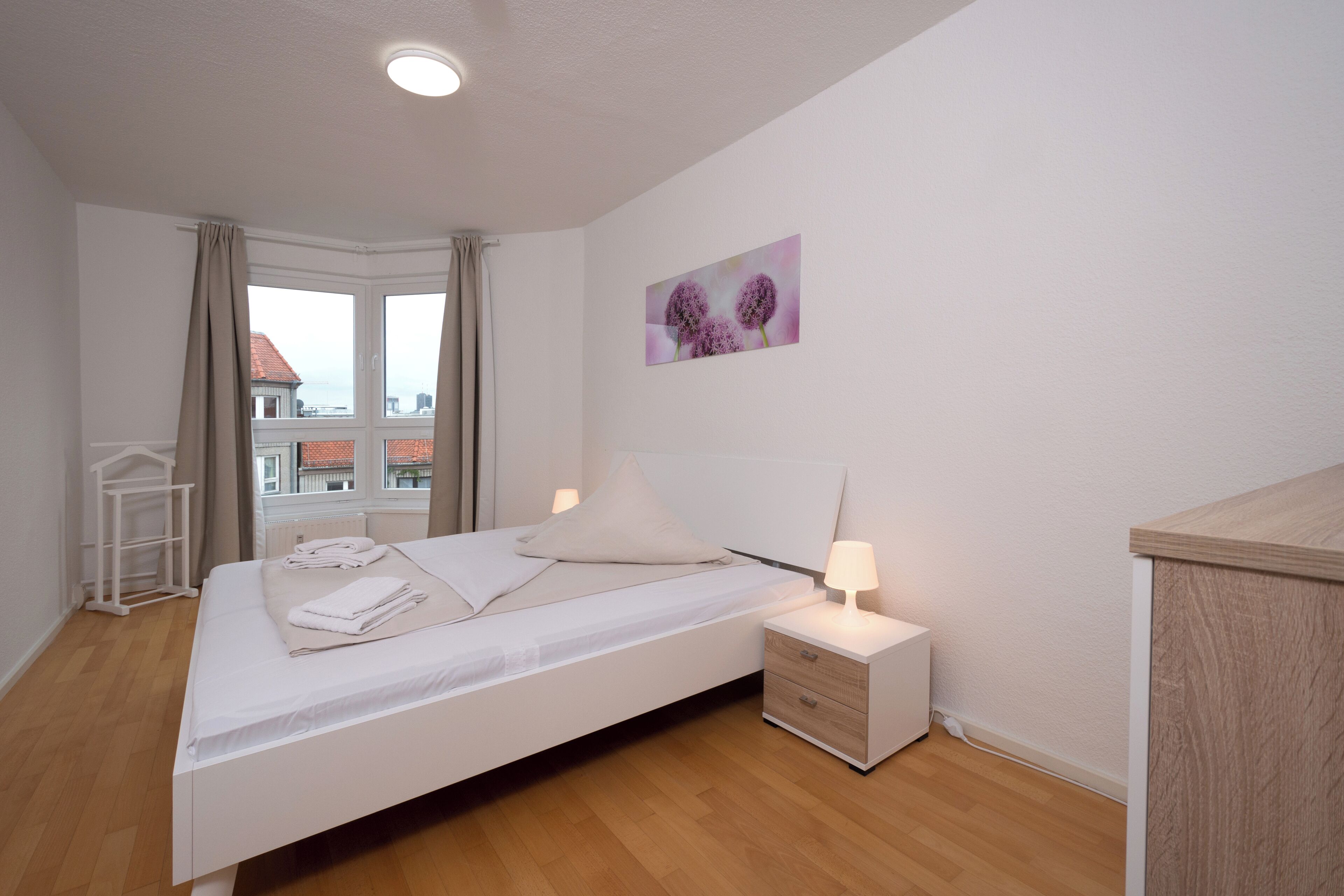 A neatly made bed with white linens, a bedside lamp, and a small bedside table with drawers. A window with curtains is visible, and a painting hangs on the wall.