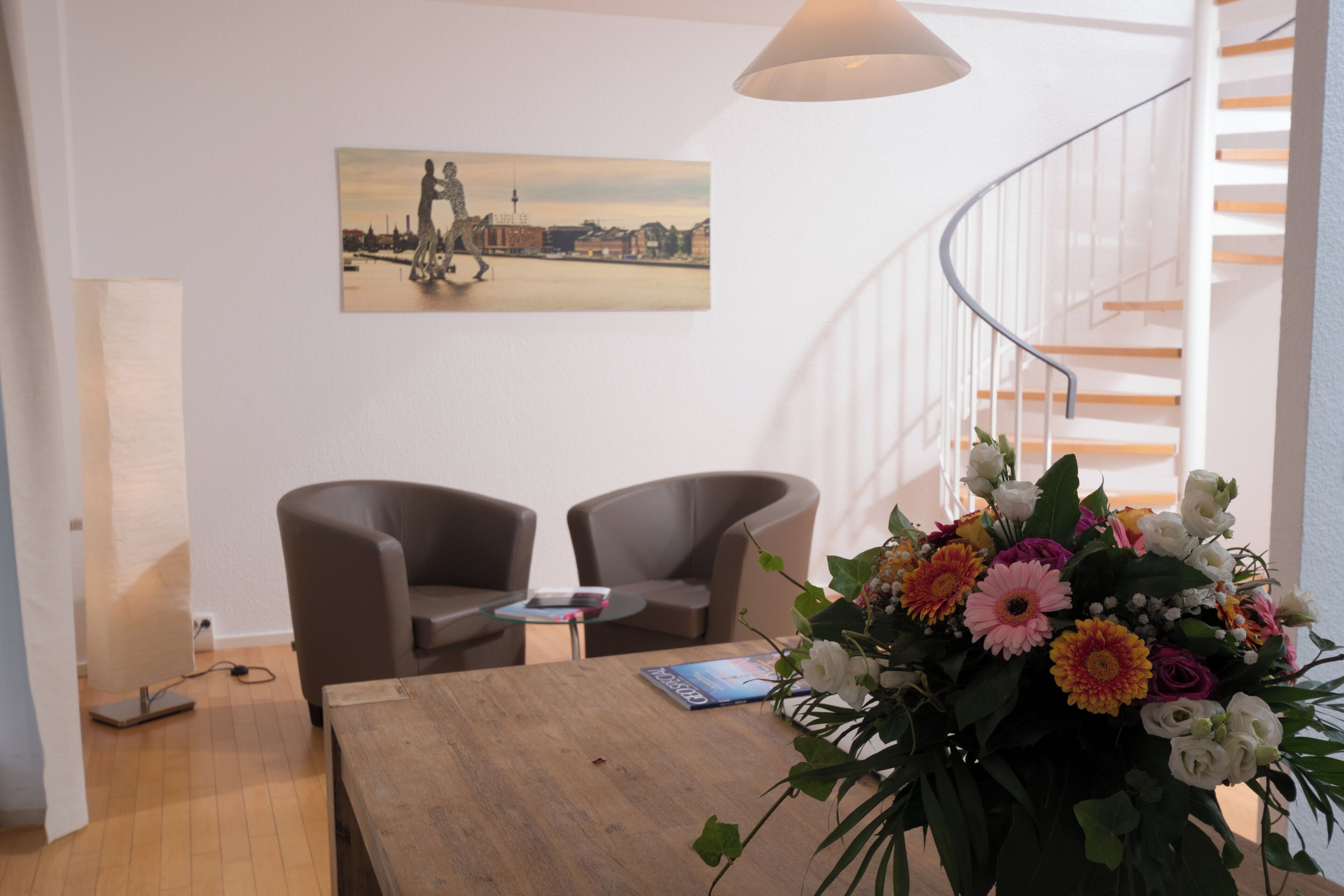 Una sala de estar moderna con una mesa de centro de madera, dos sillones, una mesa de vidrio y un arreglo floral. Un cuadro de un paisaje urbano cuelga en la pared.