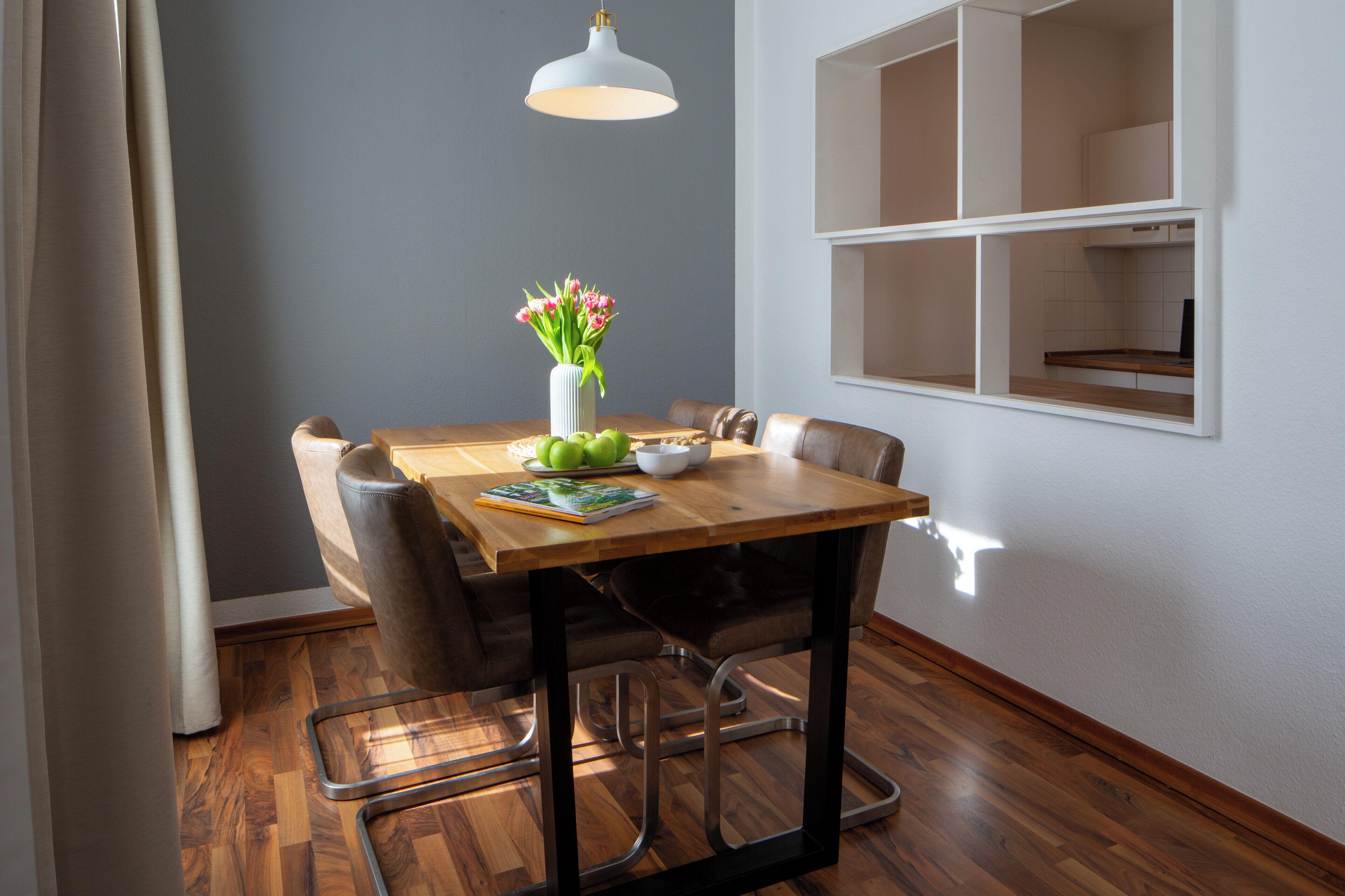 A dining area with a wooden table, chairs, a vase with flowers, and a hanging light fixture.