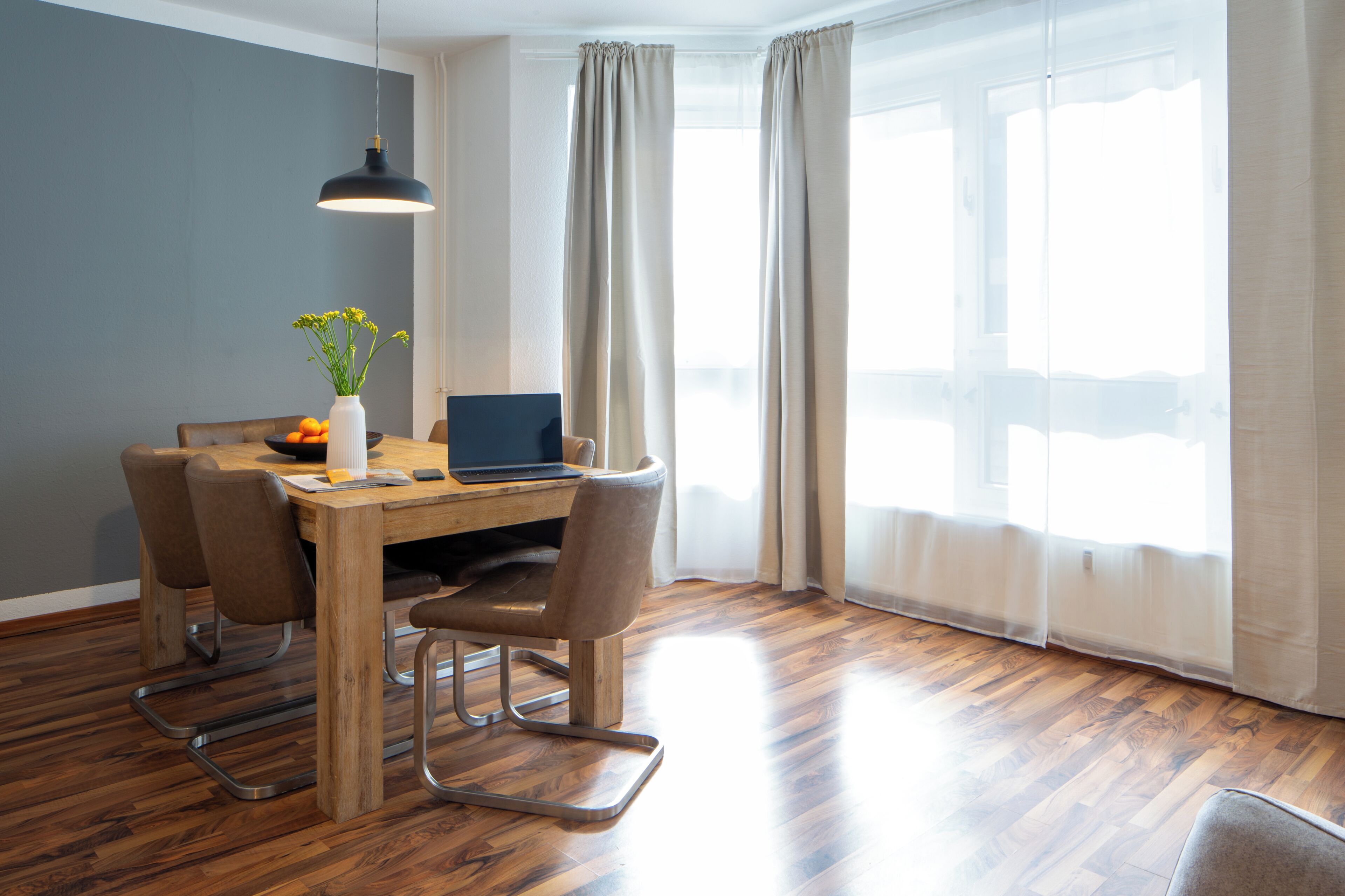 A modern dining area with a wooden table, chairs, a laptop, and a vase with flowers.
