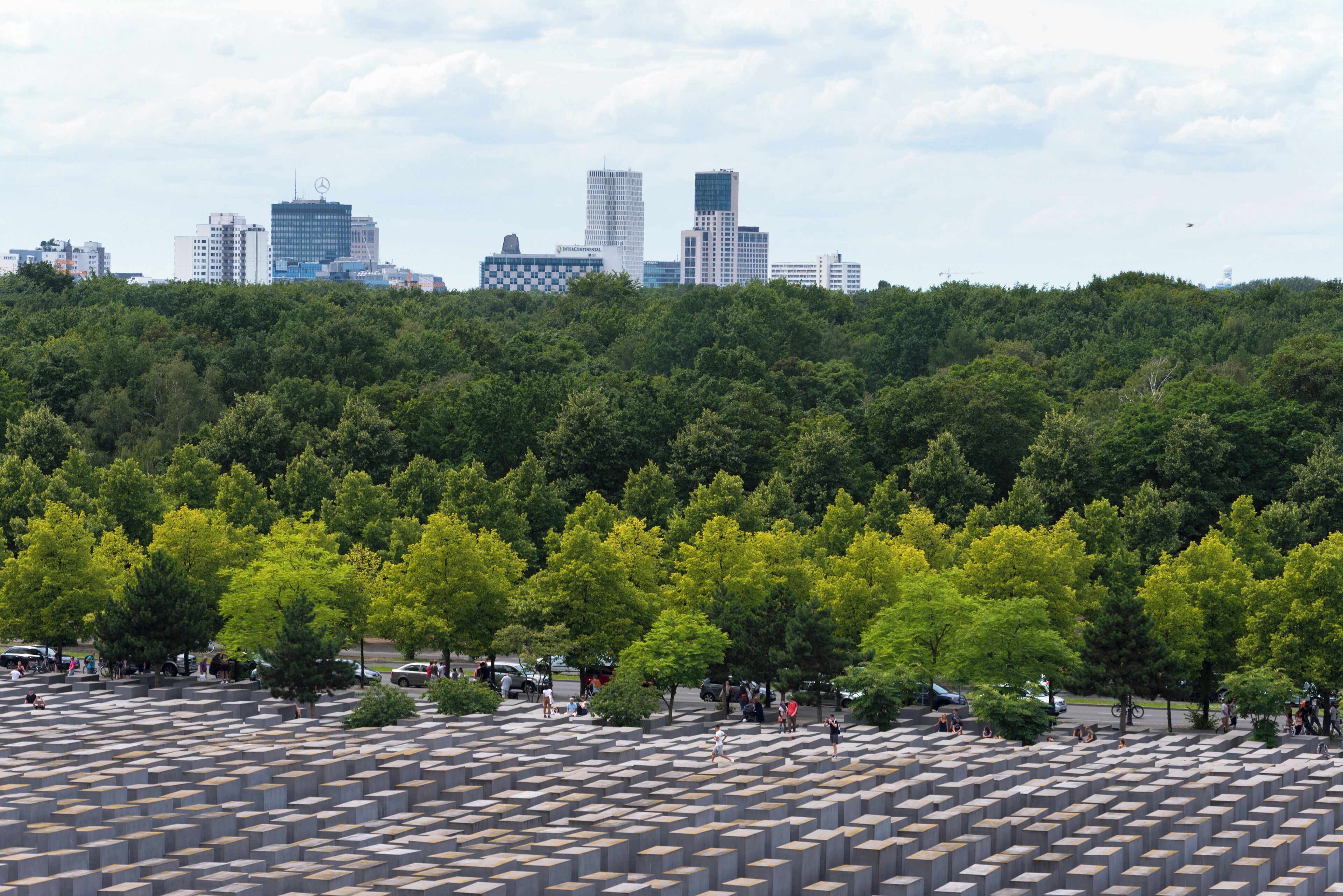 A memorial with numerous concrete blocks, surrounded by trees and a city skyline in the background.