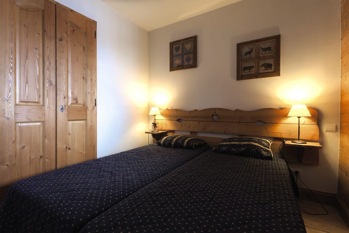 A bedroom with two beds, wooden wardrobe, wall-mounted lamps, and framed pictures.