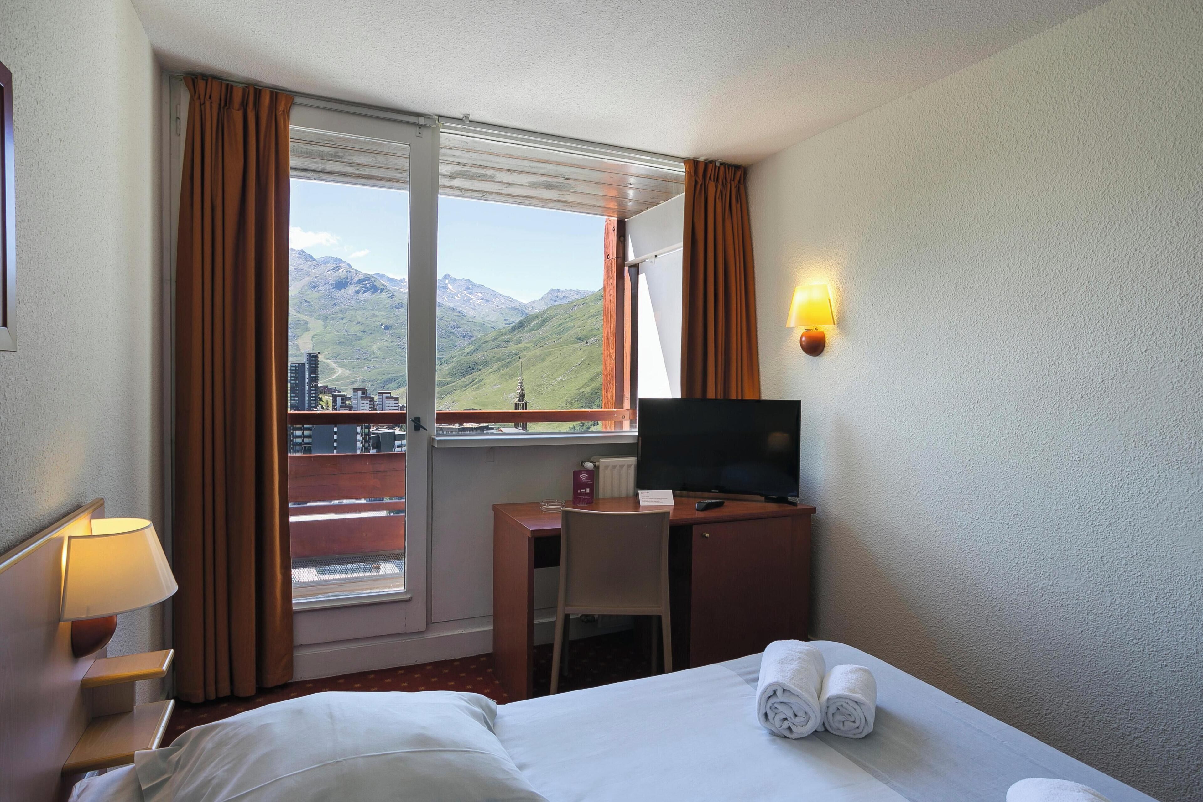 A hotel room with a bed, a desk, a TV, a lamp, a window with curtains, and a view of mountains.