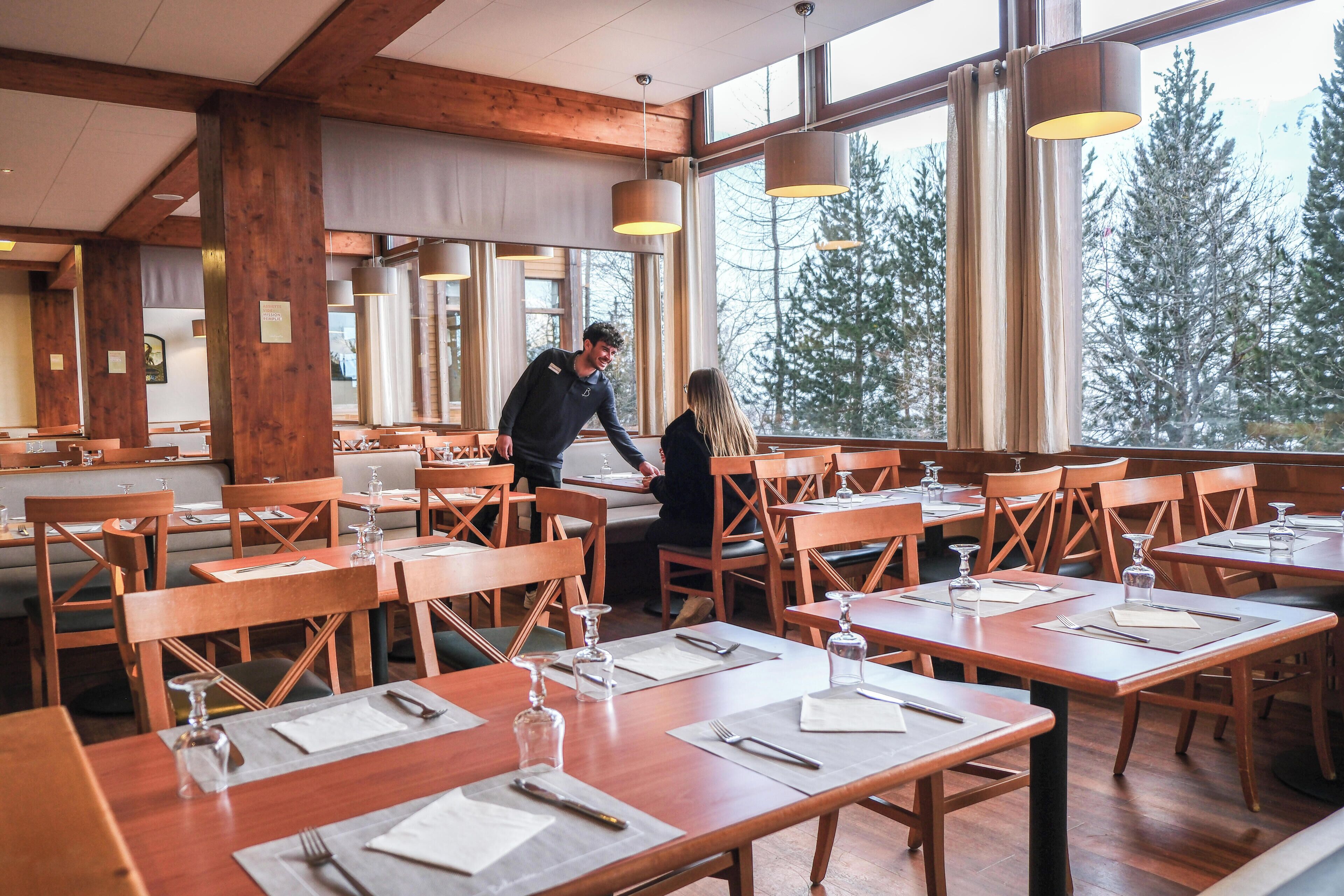 Un restaurante con mesas y sillas de madera, ventanas amplias y lámparas colgantes.