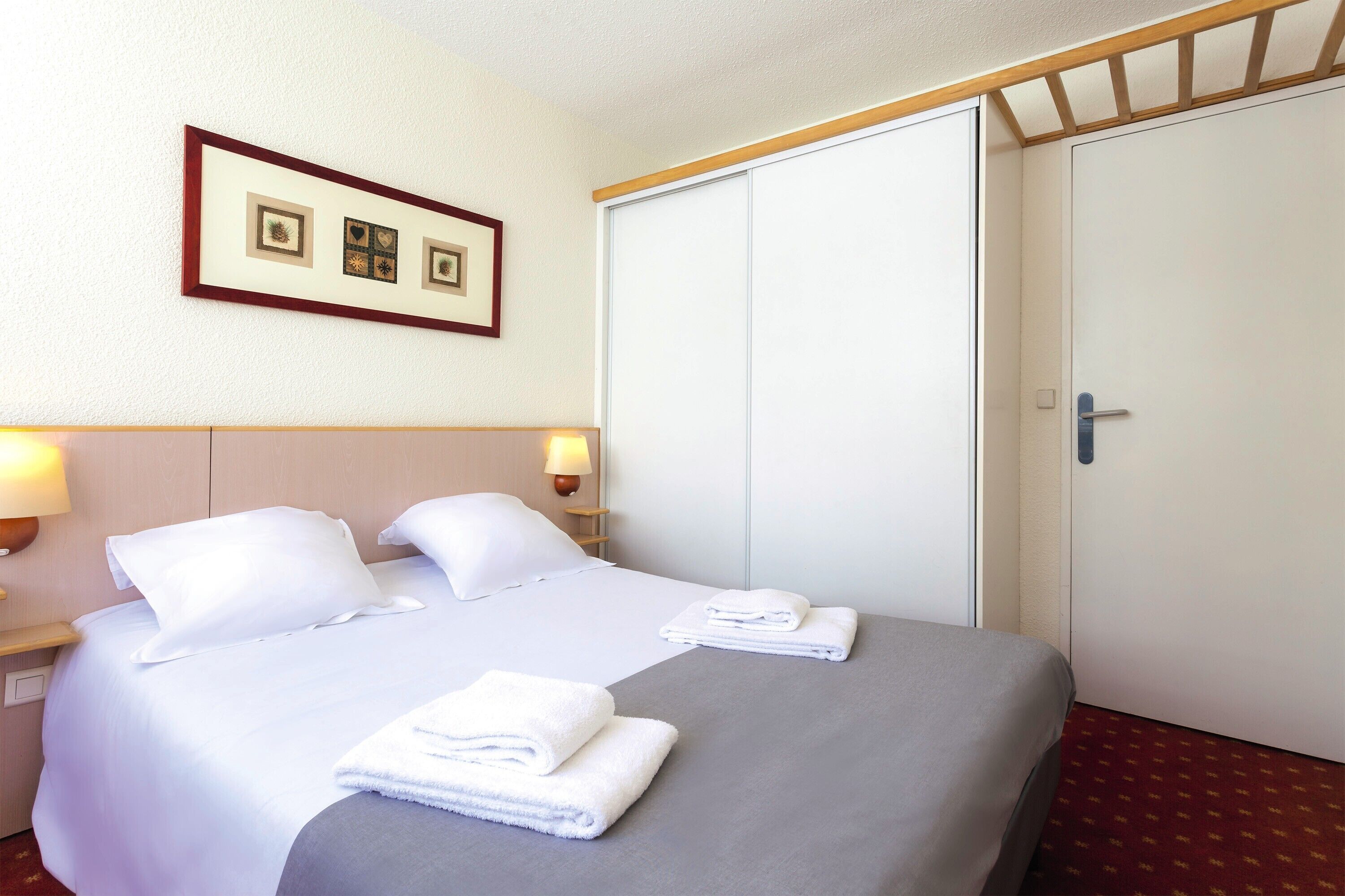 A hotel room with a bed, two pillows, a folded towel, a nightstand with a lamp, and a framed picture on the wall.