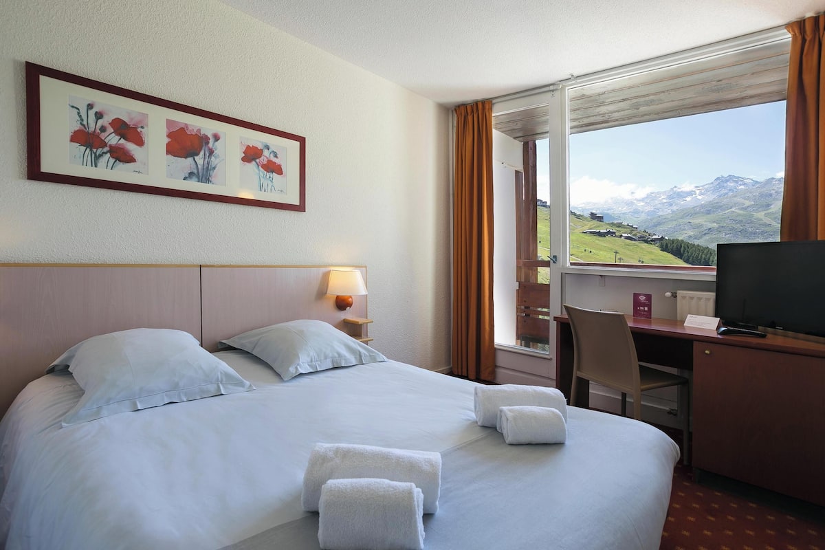 Ein Hotelzimmer mit Bett, Schreibtisch mit Fernseher, Stuhl und Fenster mit Bergblick.