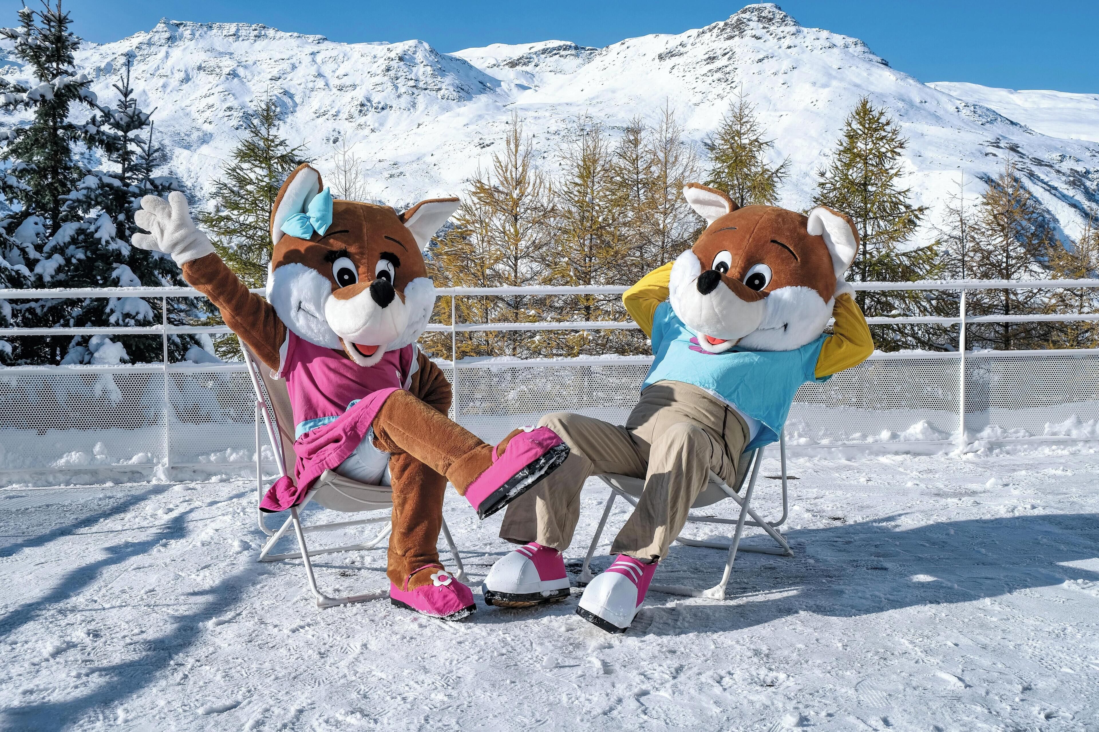 Two mascots seated on chairs in a snowy outdoor setting with a mountain backdrop.