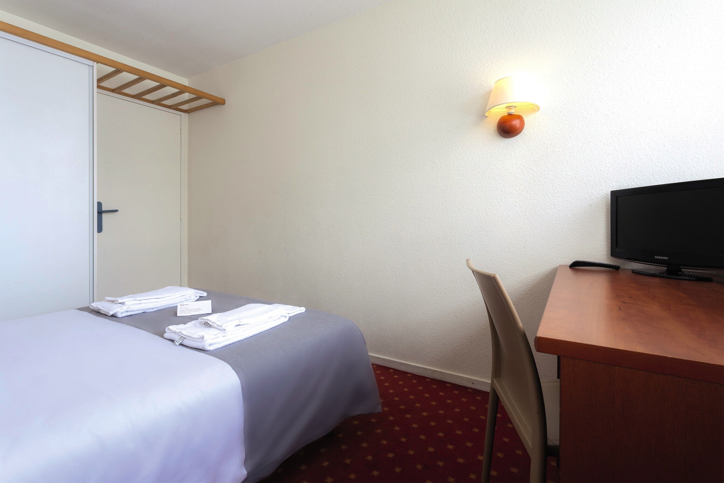 Una habitación de hotel con una cama, un escritorio con televisor y una lámpara de pared.