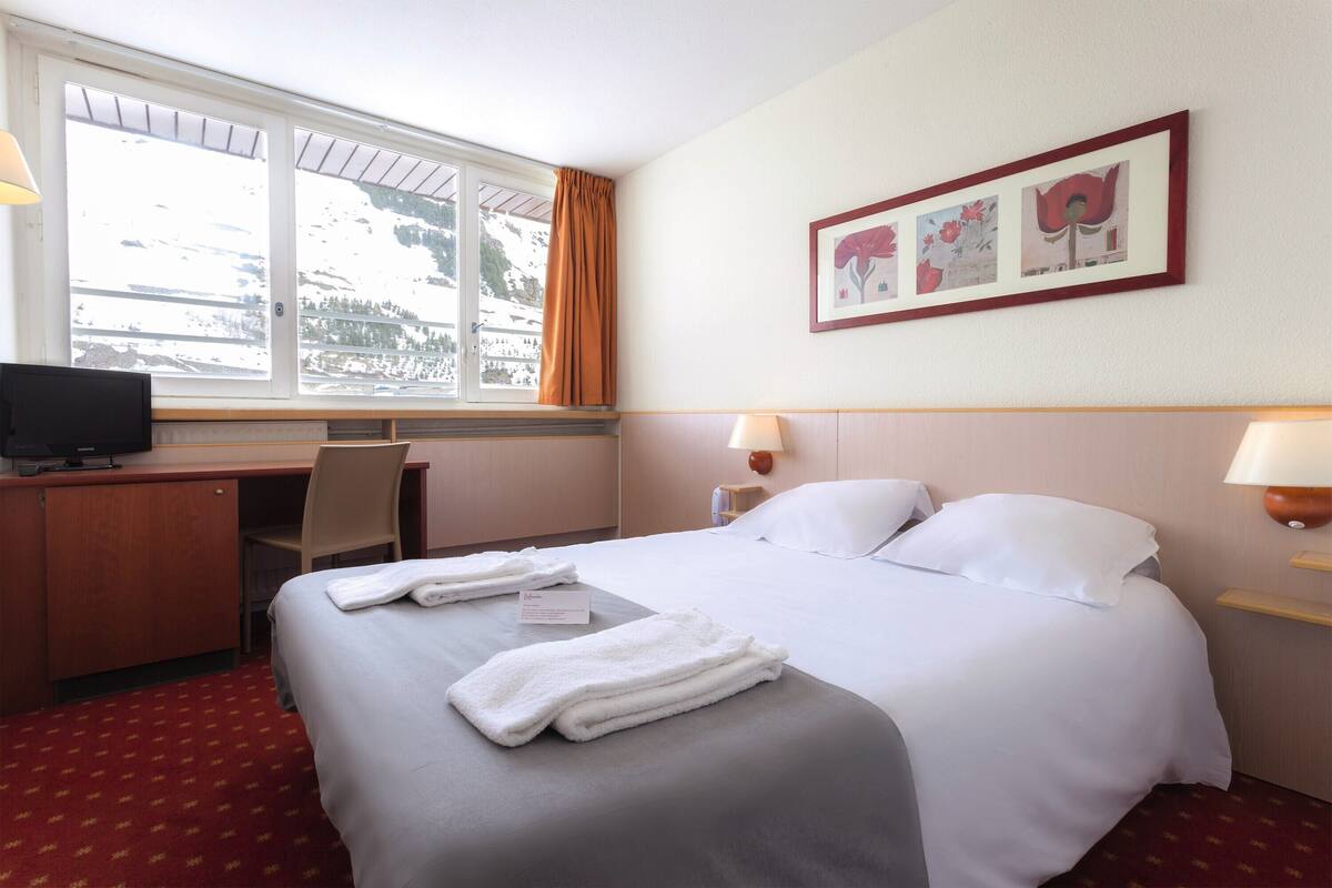 Ein Hotelzimmer mit Bett, Schreibtisch, Stuhl, Fernseher und einem Fenster mit Blick auf schneebedeckte Berge.