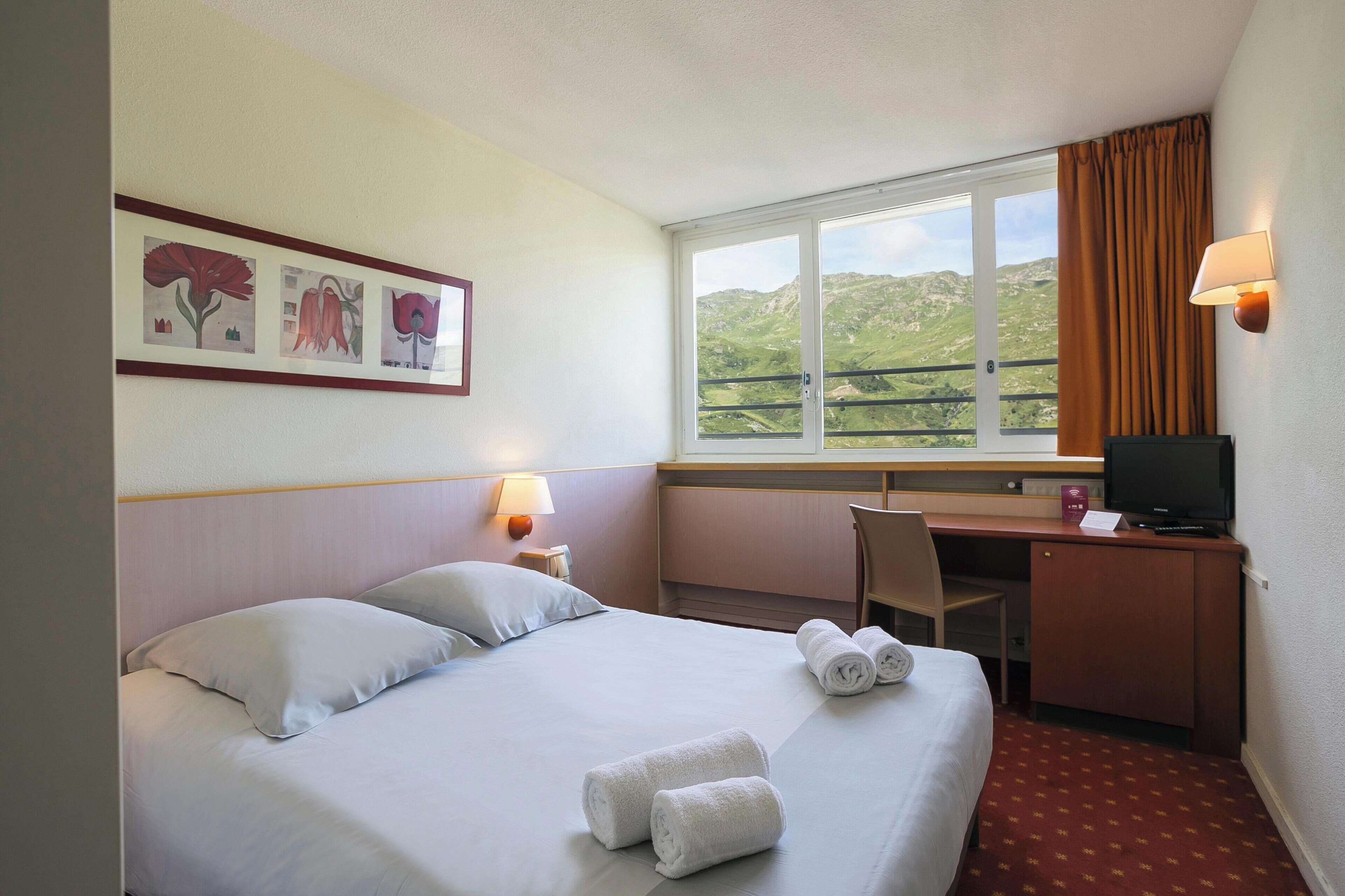 A hotel room with a bed, a desk, a chair, a television, and a window with a view of hills.
