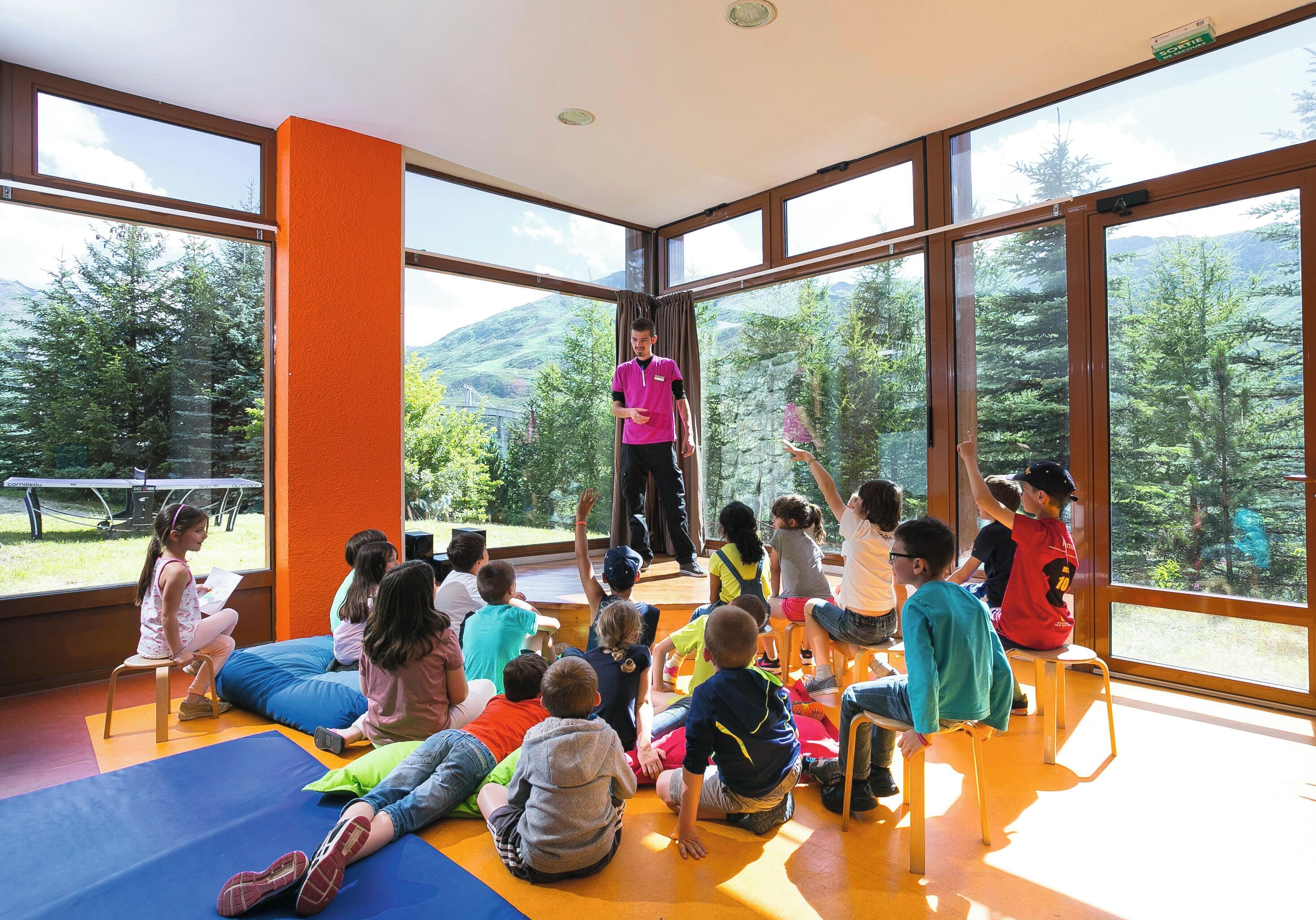 A group of children seated on mats in a room with large windows and an orange wall.