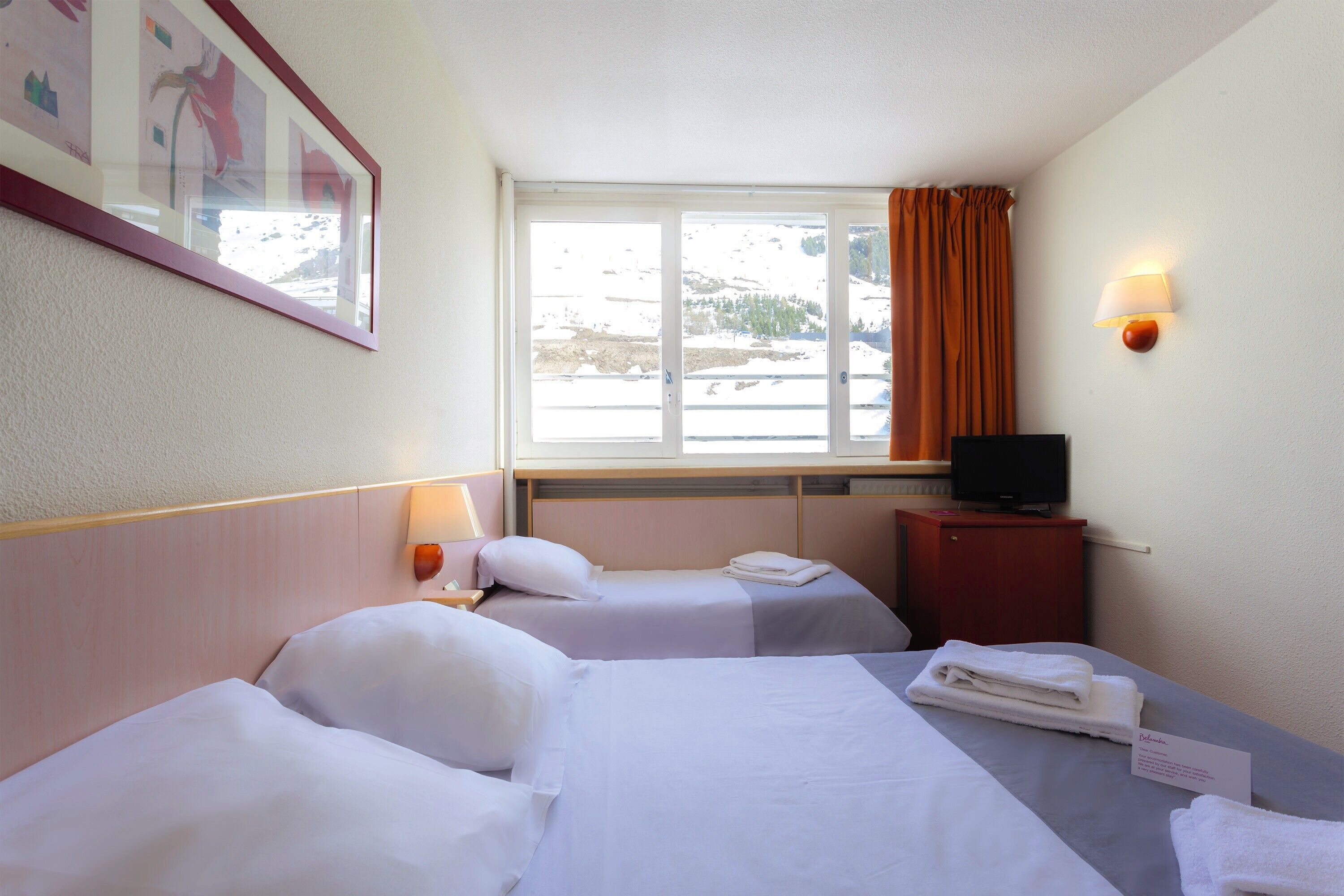Une chambre d’hôtel avec deux lits, une télévision, une fenêtre avec des rideaux et un tableau encadré au mur.