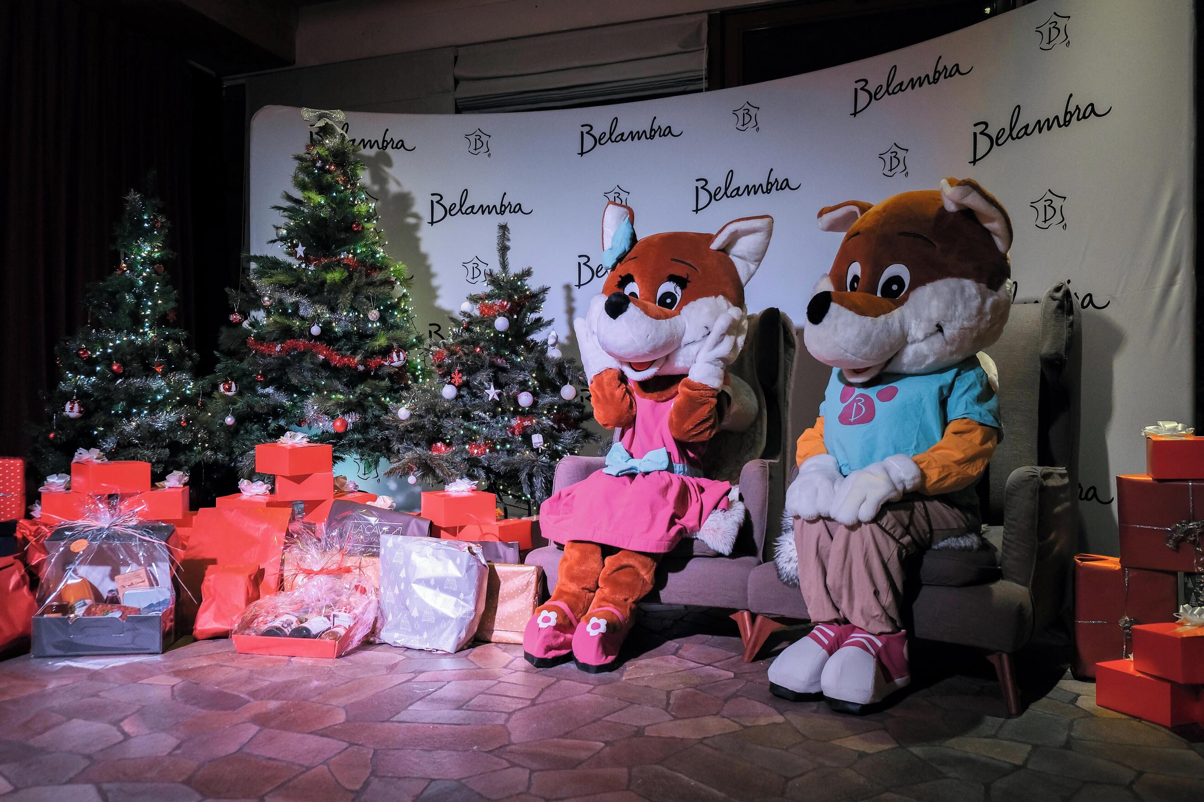 Deux mascottes sont assises devant un sapin de Noël, avec une décoration représentant le mot « Belambra » en arrière-plan.