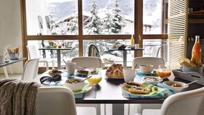 A breakfast table with various dishes, a window with a snowy mountain view, and a wooden bookshelf. - Belambra Clubs Praz-sur-Arly - L'Alisier (Praz-sur-Arly)