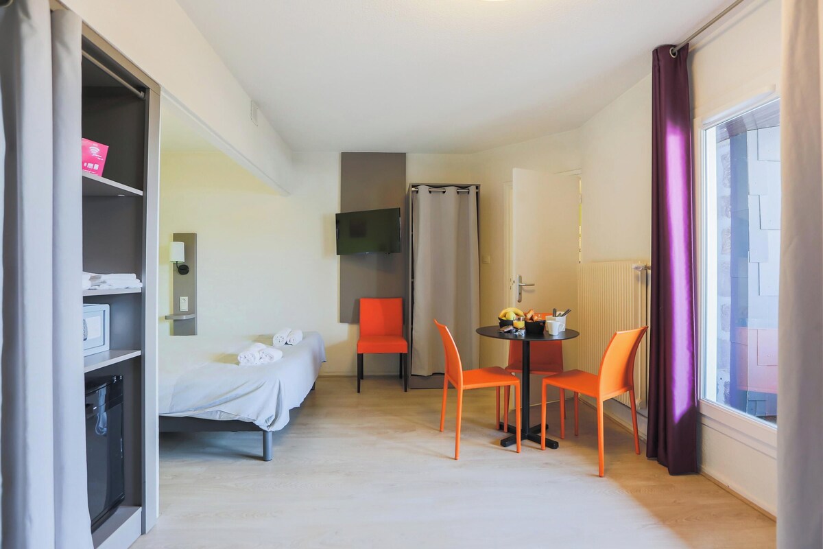 A hotel room with a bed, a desk with a chair, a television mounted on the wall, and a window with curtains.