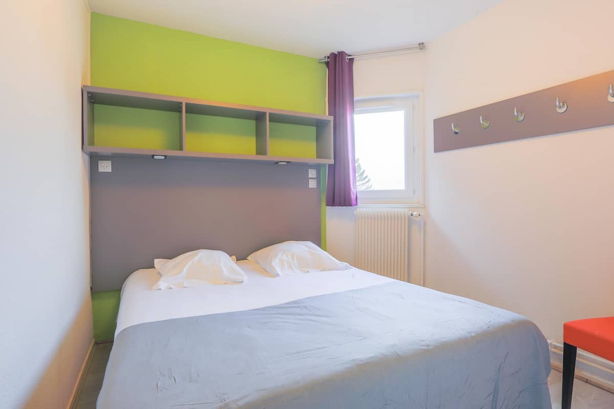 Une chambre à coucher comprenant un lit, une étagère, une fenêtre avec des rideaux et un porte-manteau fixé au mur.