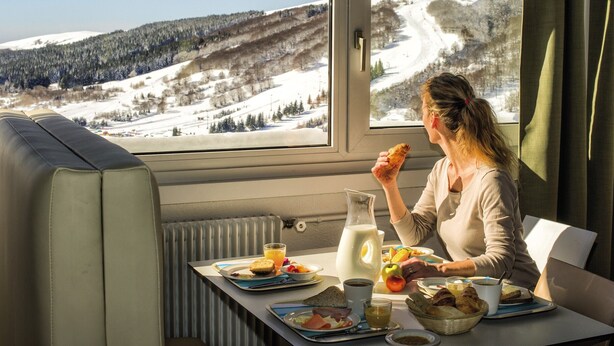 A dining area with a table set for breakfast, a window with a snowy mountain view, and a radiator under the table. - Belambra Clubs Superbesse - Le Chambourguet (Besse-et-Saint-Anastaise)