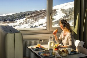 Un comedor con una mesa dispuesta para el desayuno, una ventana con vistas a una montaña nevada y un radiador debajo de la mesa.