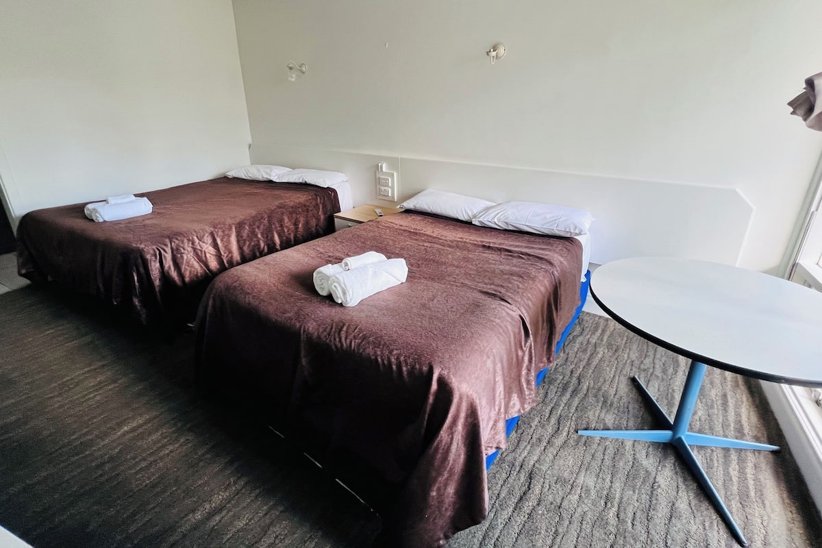 A hotel room with two beds, a small table, and a window.