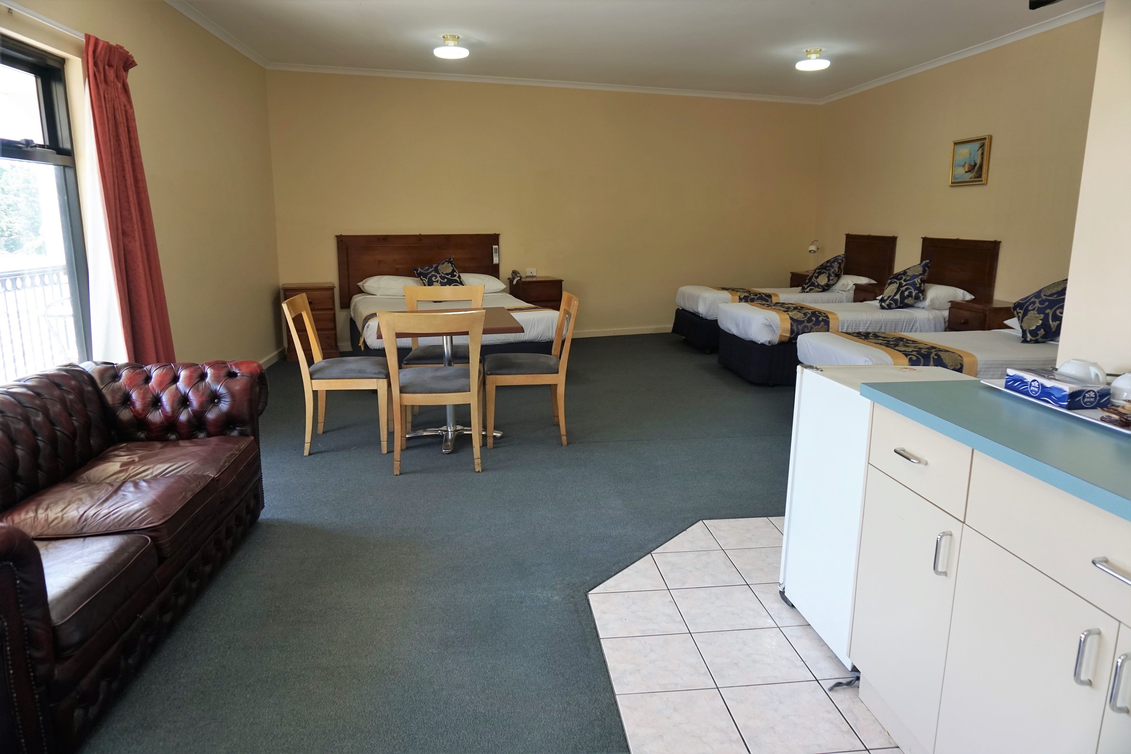 Habitación de hotel con dos camas individuales, una silla y una mesa de madera, un sofá de cuero y una kitchenette.