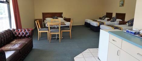 Habitación de hotel con dos camas individuales, una silla y una mesa de madera, un sofá de cuero y una kitchenette.