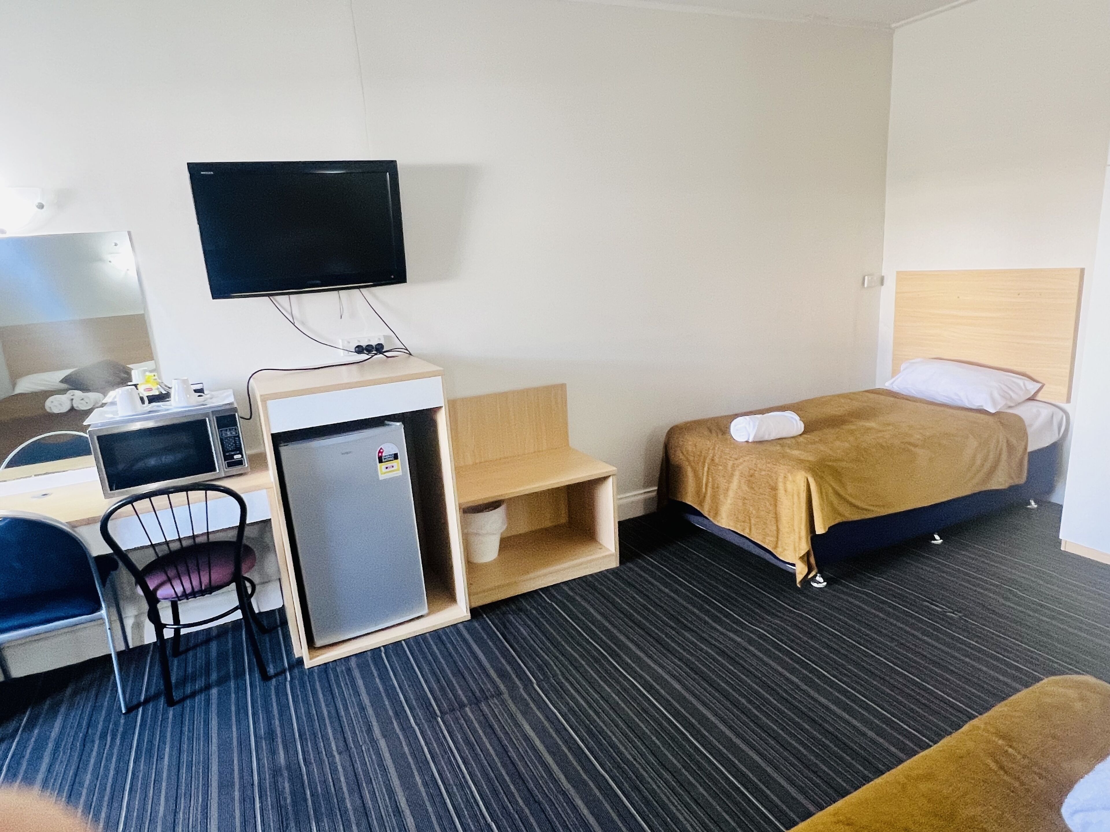 Hotellihuone, jossa on sänky, työpöytä, jossa on tietokone ja mikroaaltouuni, pieni jääkaappi, seinälle asennettu televisio ja tuoli.