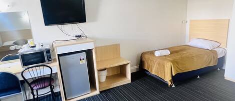 A hotel room with a bed, a desk with a computer and a microwave, a small refrigerator, a TV mounted on the wall, and a chair.