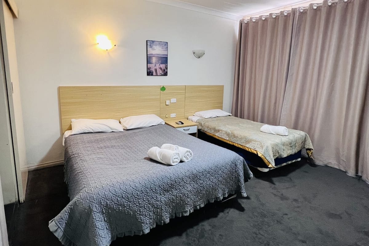 A hotel room with two beds, a headboard, a wall-mounted light, a painting, and a curtain.