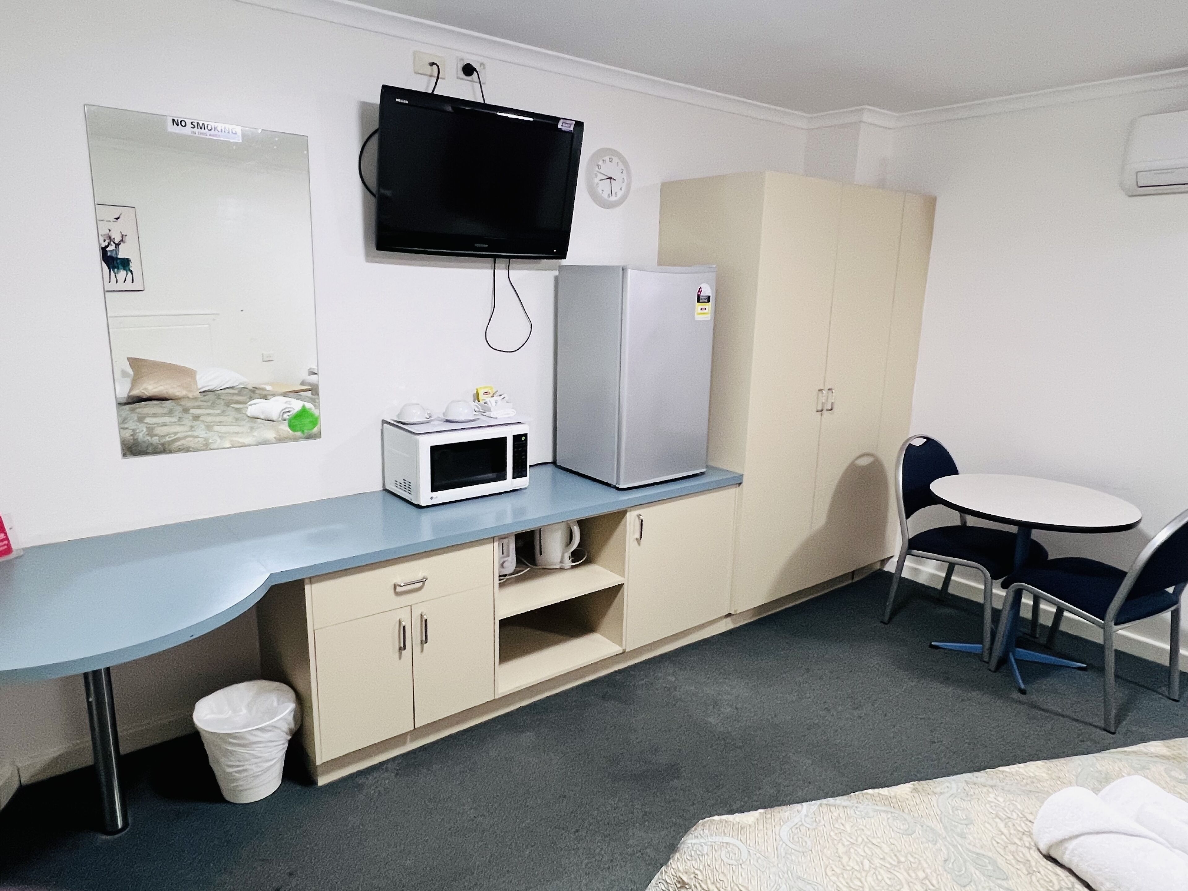 A small, compact room with a kitchenette area including a microwave, refrigerator, and a small table with two chairs. A television is mounted on the wall above the kitchenette.