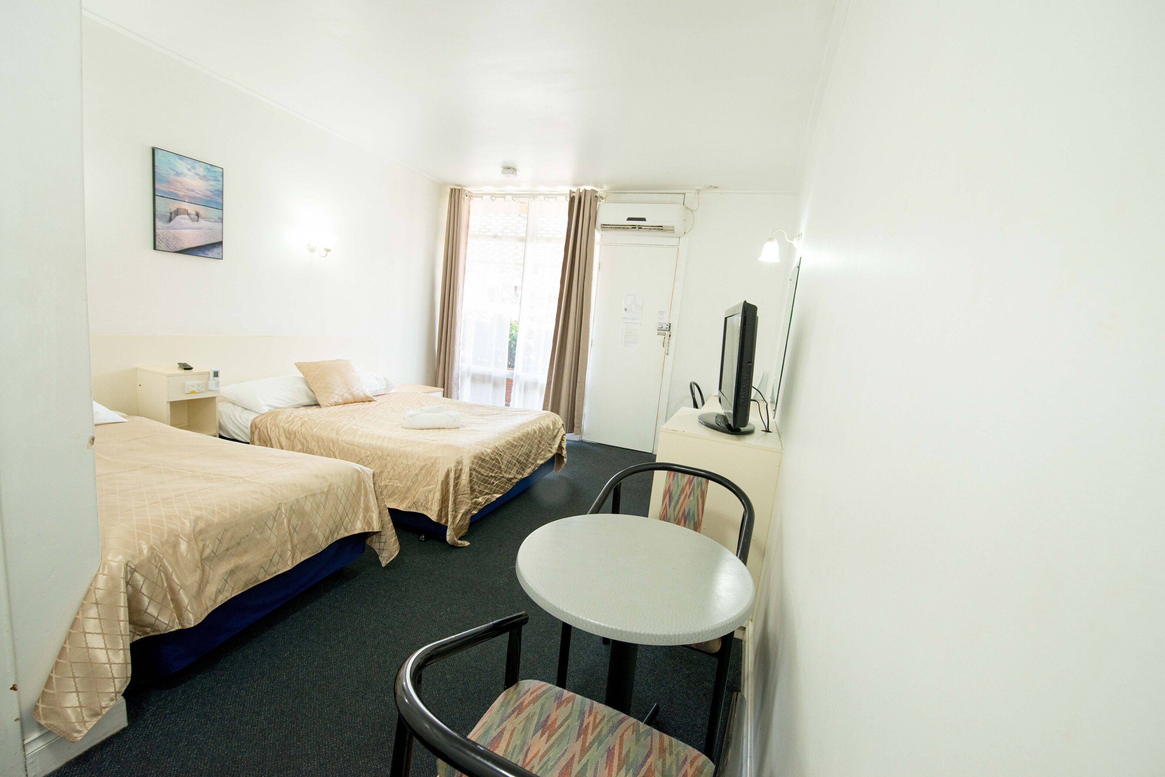 A hotel room with two beds, a small round table, a chair, a TV, and a window with curtains.