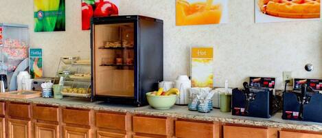 Una barra de desayuno con refrigerador, un bol de fruta y un cartel que indica bebidas calientes.