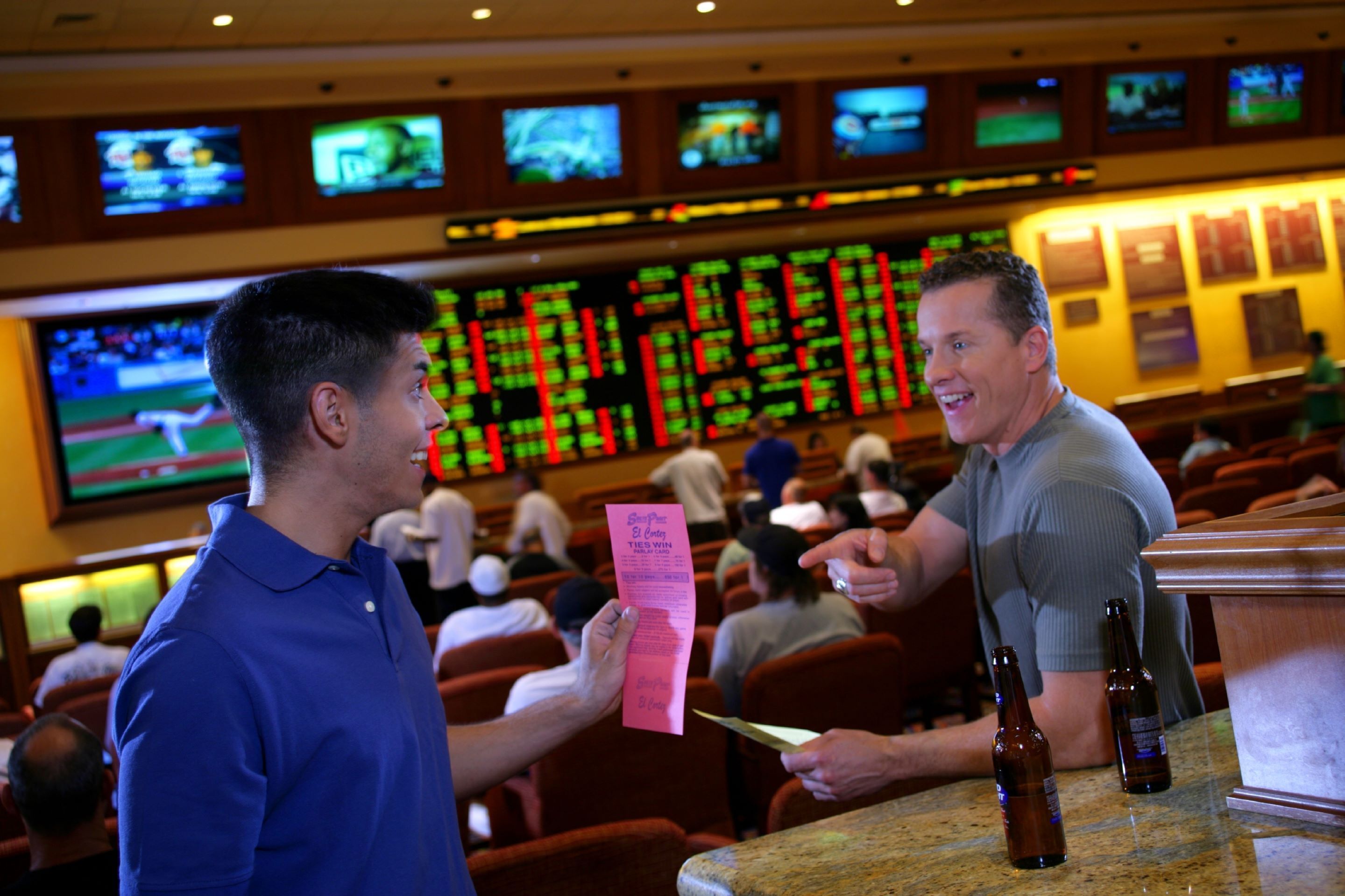 Un hombre sostiene un boleto rosa en un casino, con múltiples pantallas mostrando información al fondo.