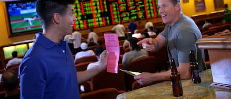Un hombre sostiene un boleto rosa en un casino, con múltiples pantallas mostrando información al fondo.