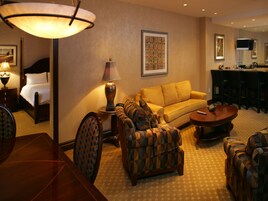 Una habitación de hotel con cama, sofá, mesita de café, sillón estampado, lámpara y un cuadro en la pared.