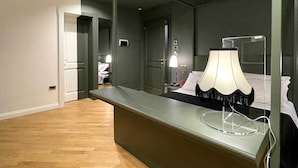 Una habitación de hotel con un espejo grande, una mesita de noche con lámpara, una cama y una puerta que da a otra habitación.