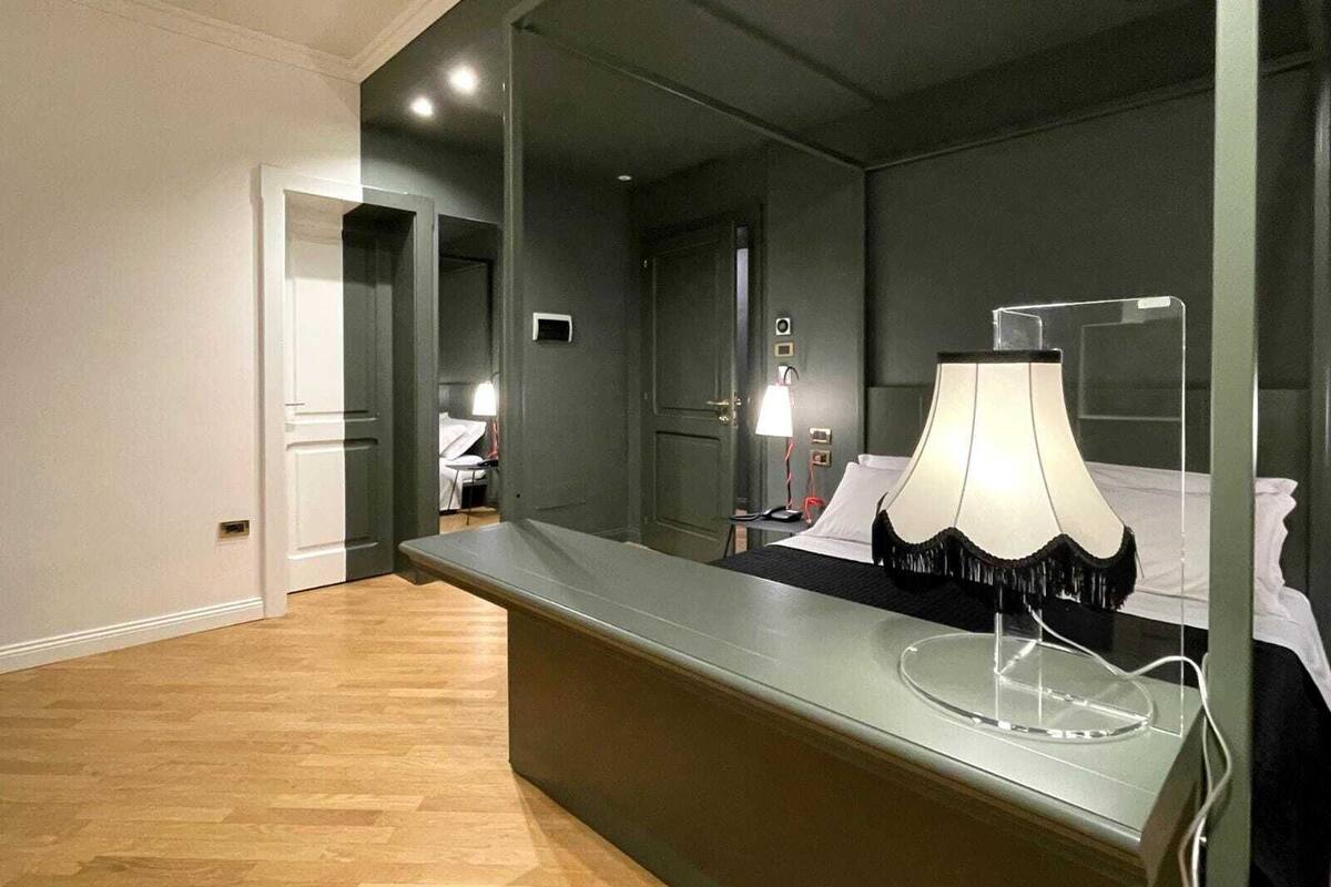 Una habitación de hotel con un espejo grande, una mesita de noche con lámpara, una cama y una puerta que da a otra habitación.