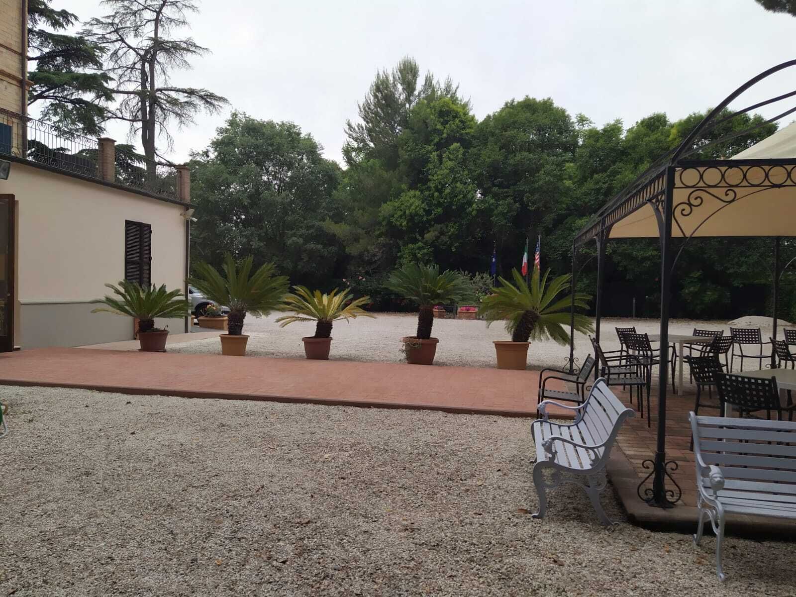 A covered outdoor seating area with tables and benches, surrounded by potted plants and trees.