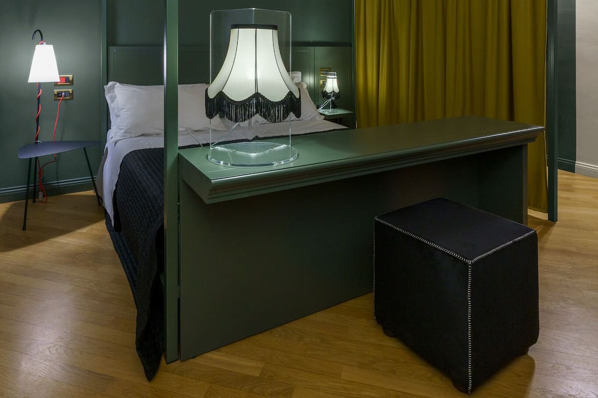 Una habitación con cama, escritorio con lámpara, un taburete pequeño y cortina.