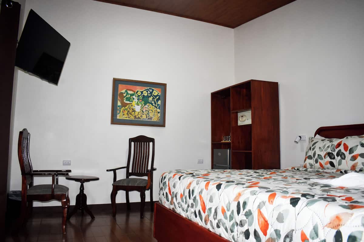 A hotel room with a bed, a desk, two chairs, a television, and a painting on the wall.