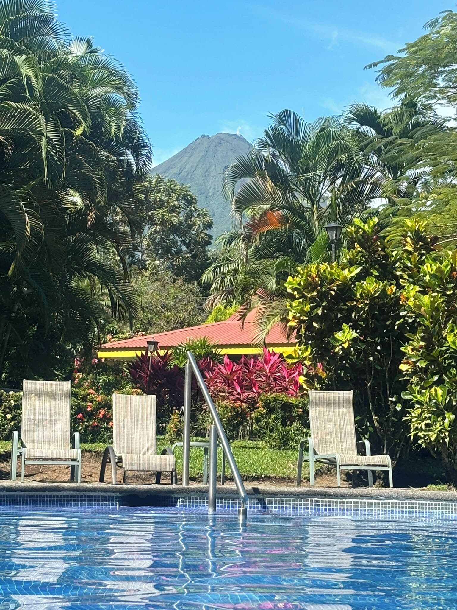 Ein Swimmingpool mit Liegestühlen, umgeben von tropischer Vegetation und einem Berg im Hintergrund.