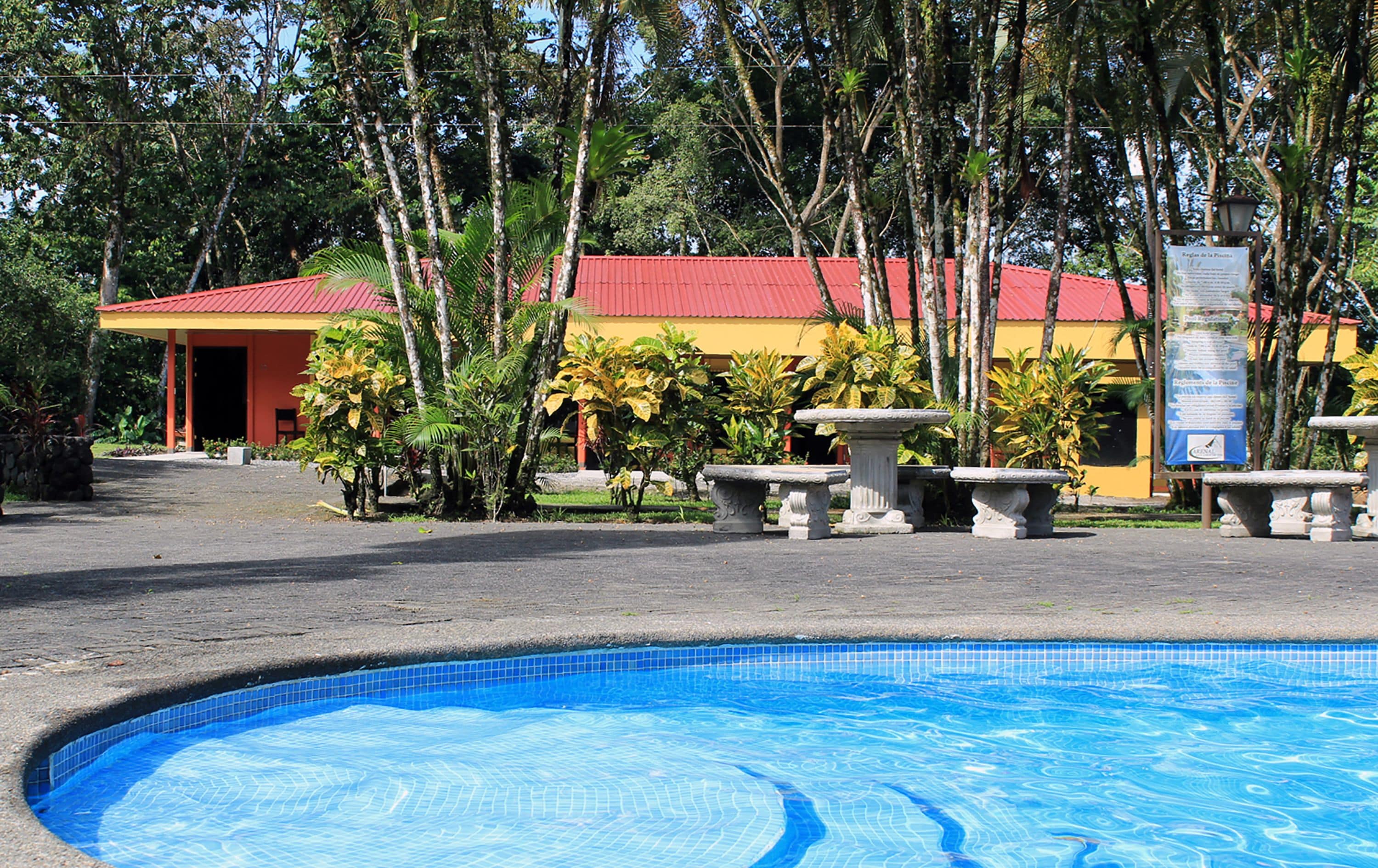 A swimming pool with a tiled edge, surrounded by a paved area with stone planters. In the background, there is a building with a red roof and yellow walls, and a signboard is visible.