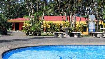 A swimming pool with a tiled edge, surrounded by a paved area with stone planters. In the background, there is a building with a red roof and yellow walls, and a signboard is visible.