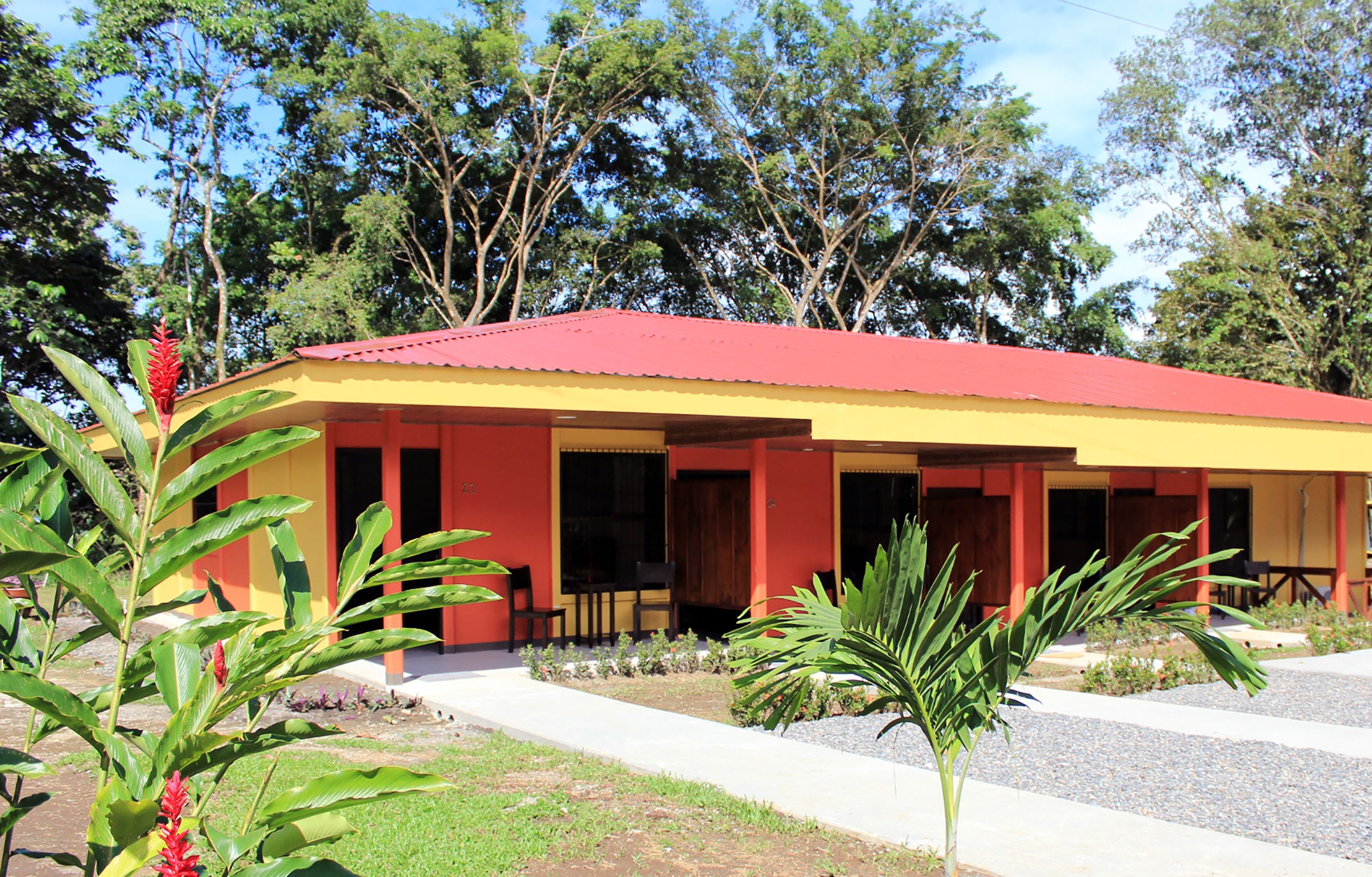 A building with a red roof and yellow walls, surrounded by greenery and trees.