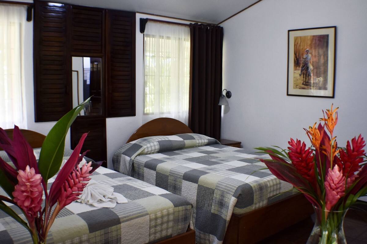 A hotel room with two beds, a window with curtains, a painting on the wall, and a vase with flowers.