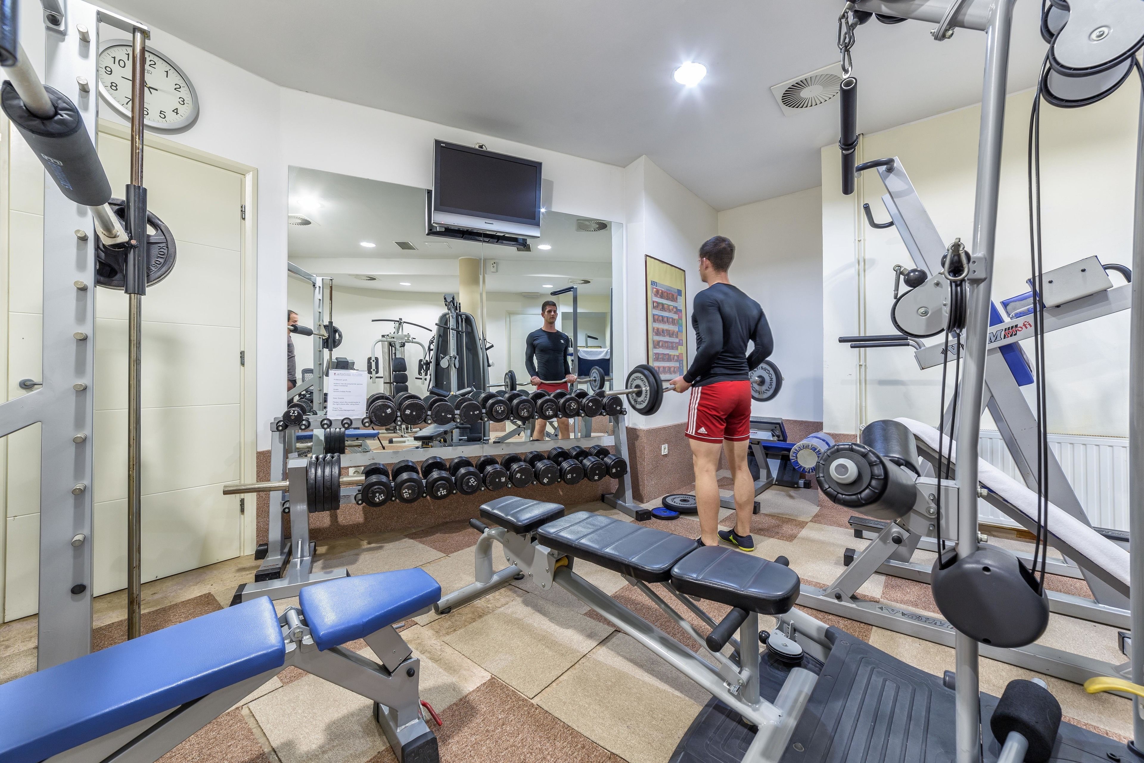 A gym with weightlifting equipment, benches, and a television on the wall.