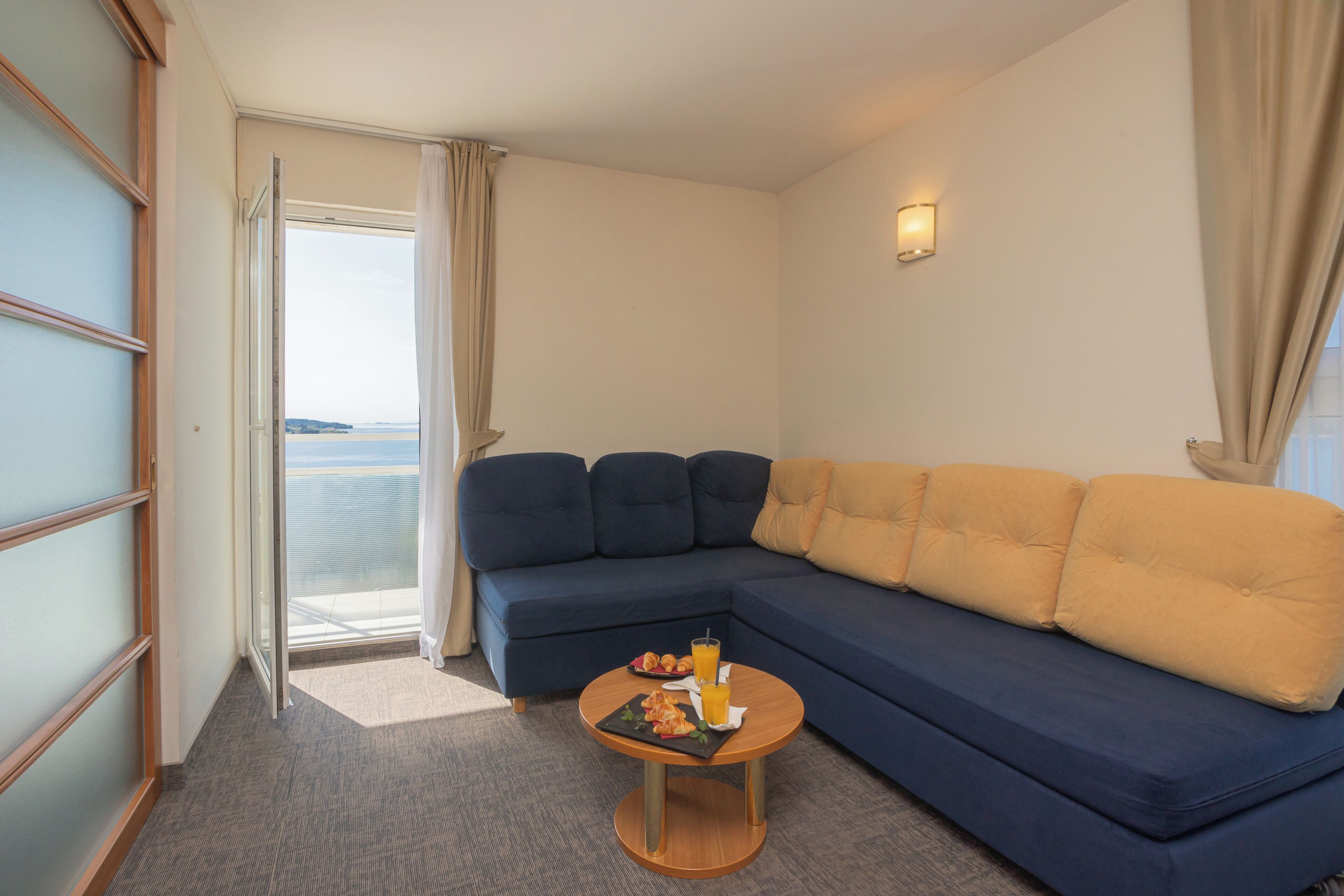 Una habitación con sofá, una mesa pequeña con comida, una ventana con cortinas y vista al mar.
